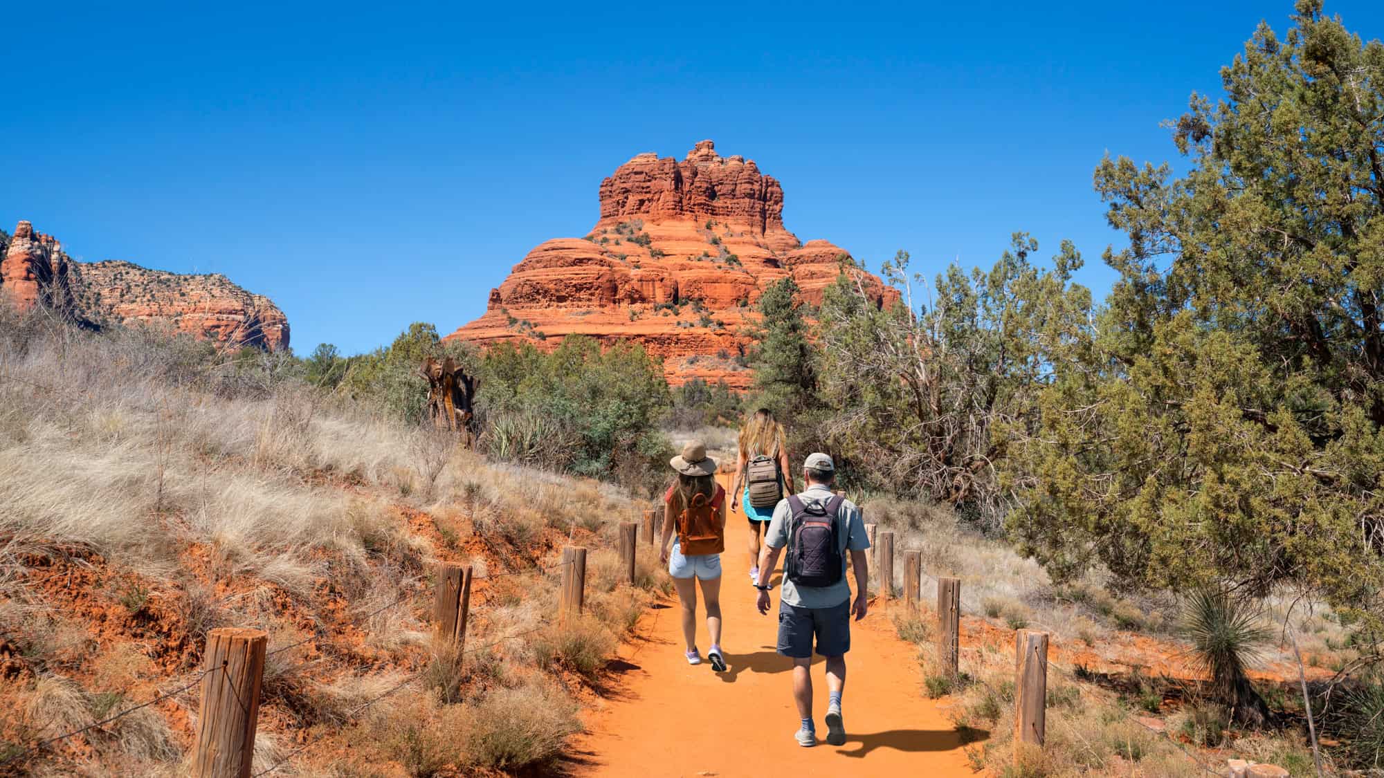 A group of hikers walks a dirt trail flanked by trees and dry grass toward the vibrant red, rounded formation of Bell Rock beneath a cloudless blue sky.
