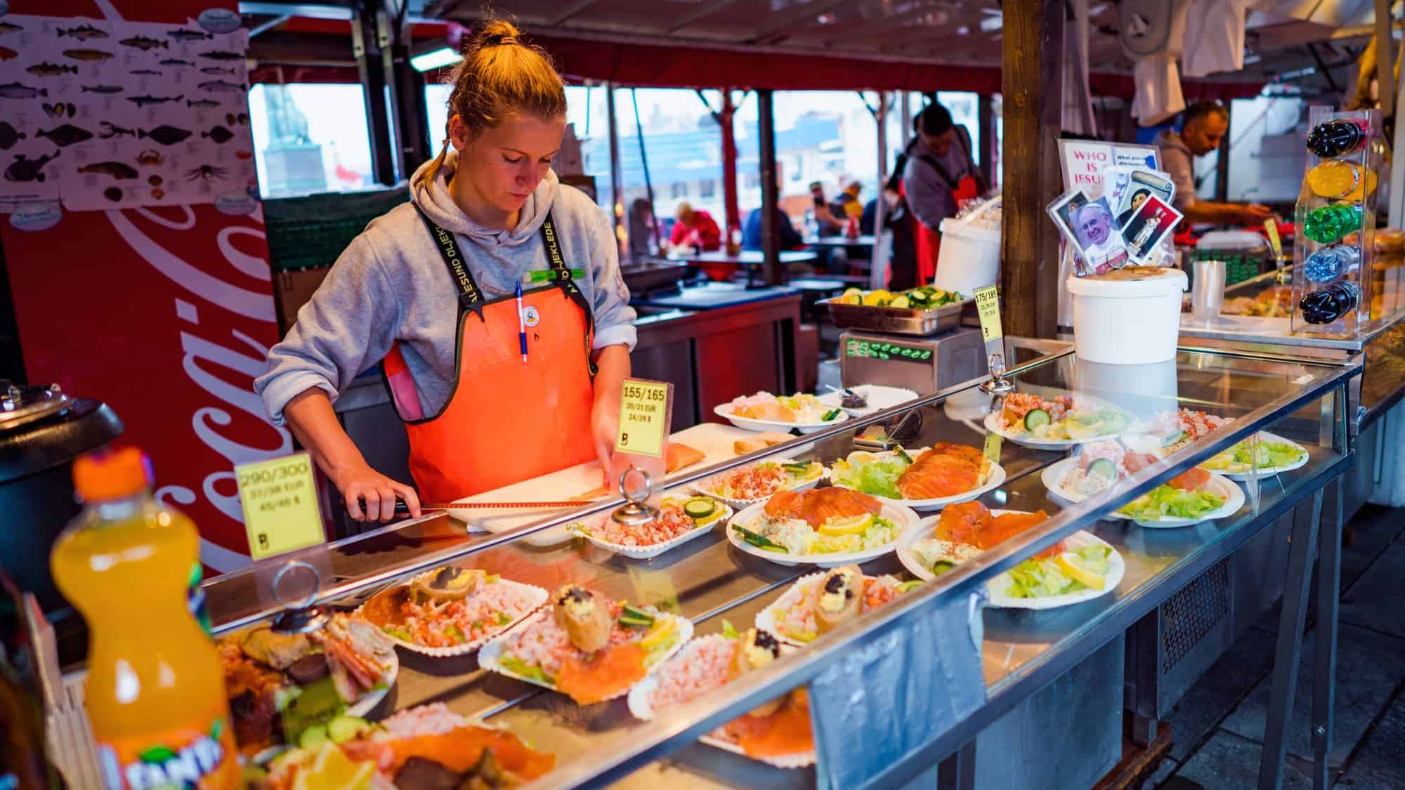 A vendor in a bright orange apron prepares seafood dishes at a bustling fish market stall, with plates of smoked salmon, shrimp, and salad displayed in a chilled glass case.