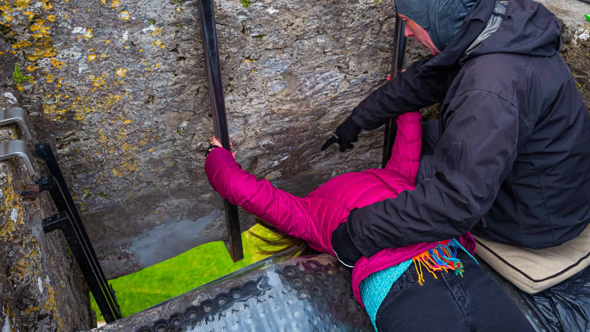 A person in a magenta jacket leans backward while gripping metal bars to kiss the Blarney Stone at Blarney Castle, with an attendant assisting. The ancient stone wall and vivid green grass below highlight this quirky Irish tradition.