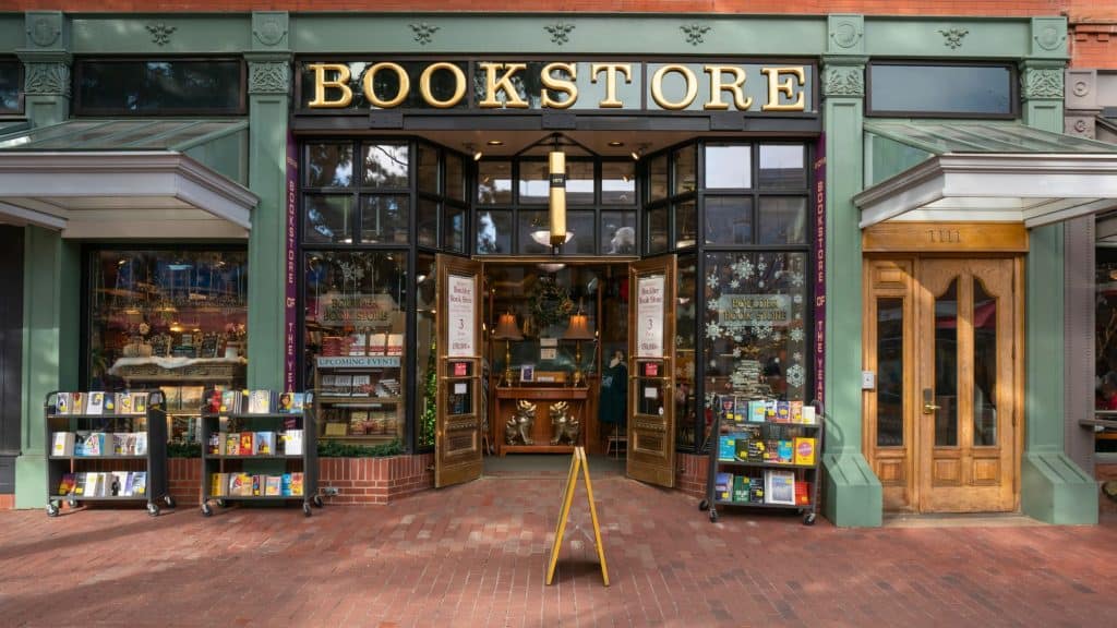 Street view of a charming bookstore entrance with large windows, racks of books outside, and signage reading “BOOKSTORE” above a welcoming double-door entrance.