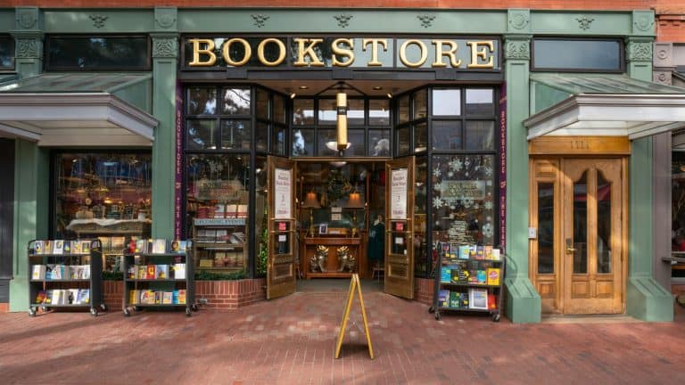 Street view of a charming bookstore entrance with large windows, racks of books outside, and signage reading “BOOKSTORE” above a welcoming double-door entrance.