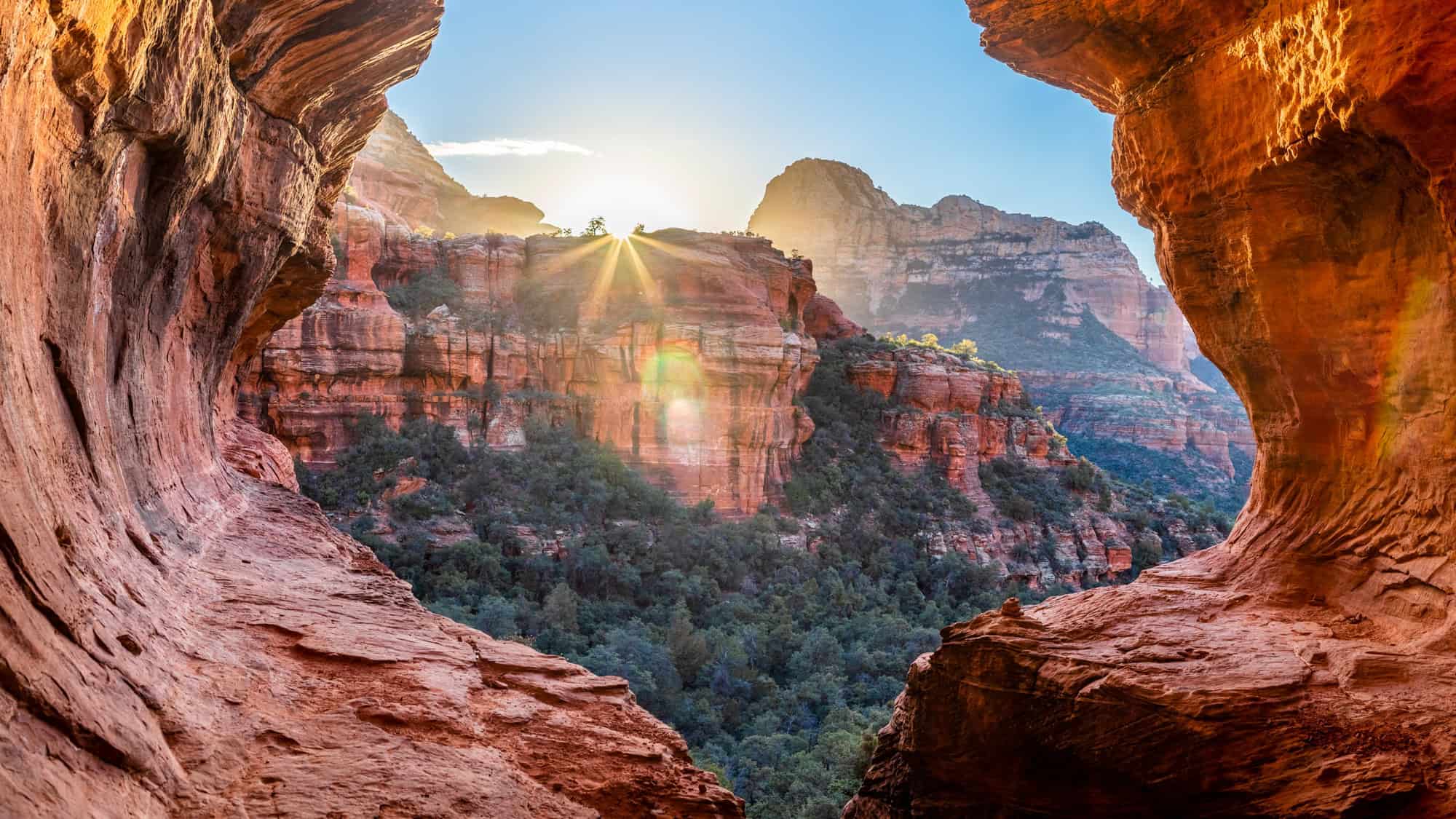 Sunlight streams through a rocky opening, illuminating red canyon walls and a lush forested valley below, capturing the dramatic beauty of Boynton Canyon in Sedona.