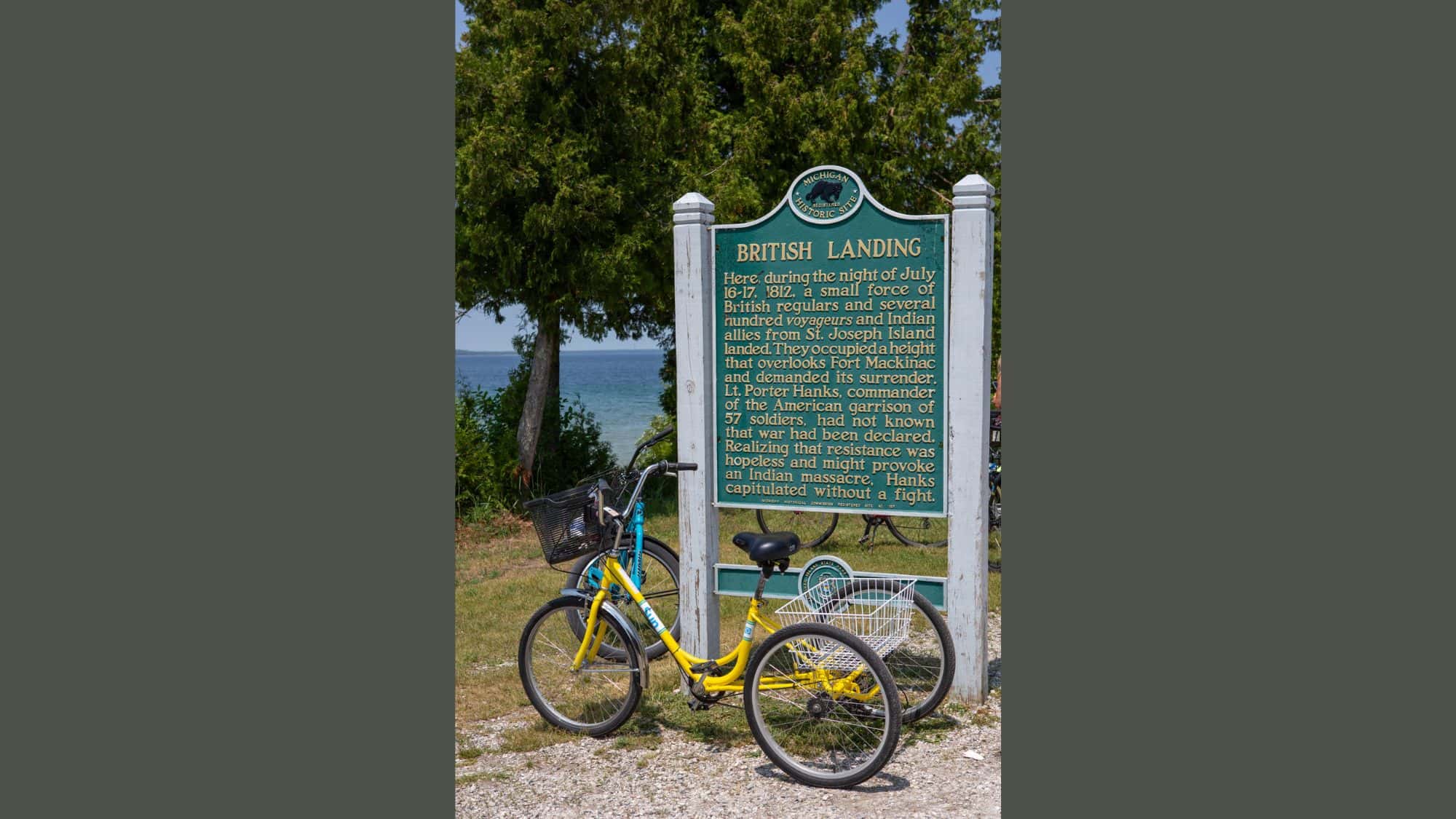 A green Michigan historic marker titled "British Landing" stands beside a yellow rental bike, with text describing the 1812 British landing during the War of 1812 and a glimpse of Lake Huron in the background.