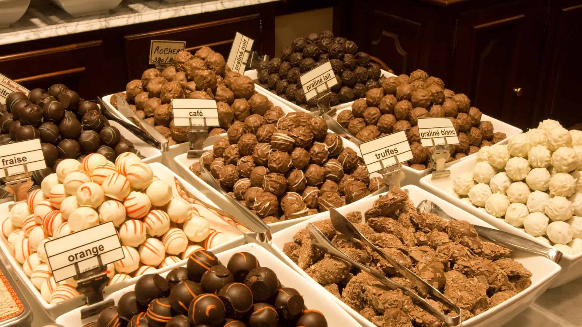 A luxurious selection of assorted Belgian chocolate truffles and pralines displayed in trays with handwritten labels in French, showcasing a variety of flavors including “fraise,” “café,” and “truffes.”