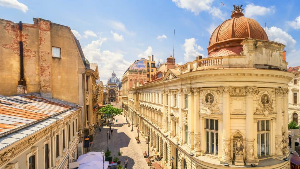 A perspective of Bucharest's Old Town district featuring grand neoclassical architecture with ornate carvings, domes, and columns, viewed from a narrow cobblestone street bustling with pedestrians and cafes.