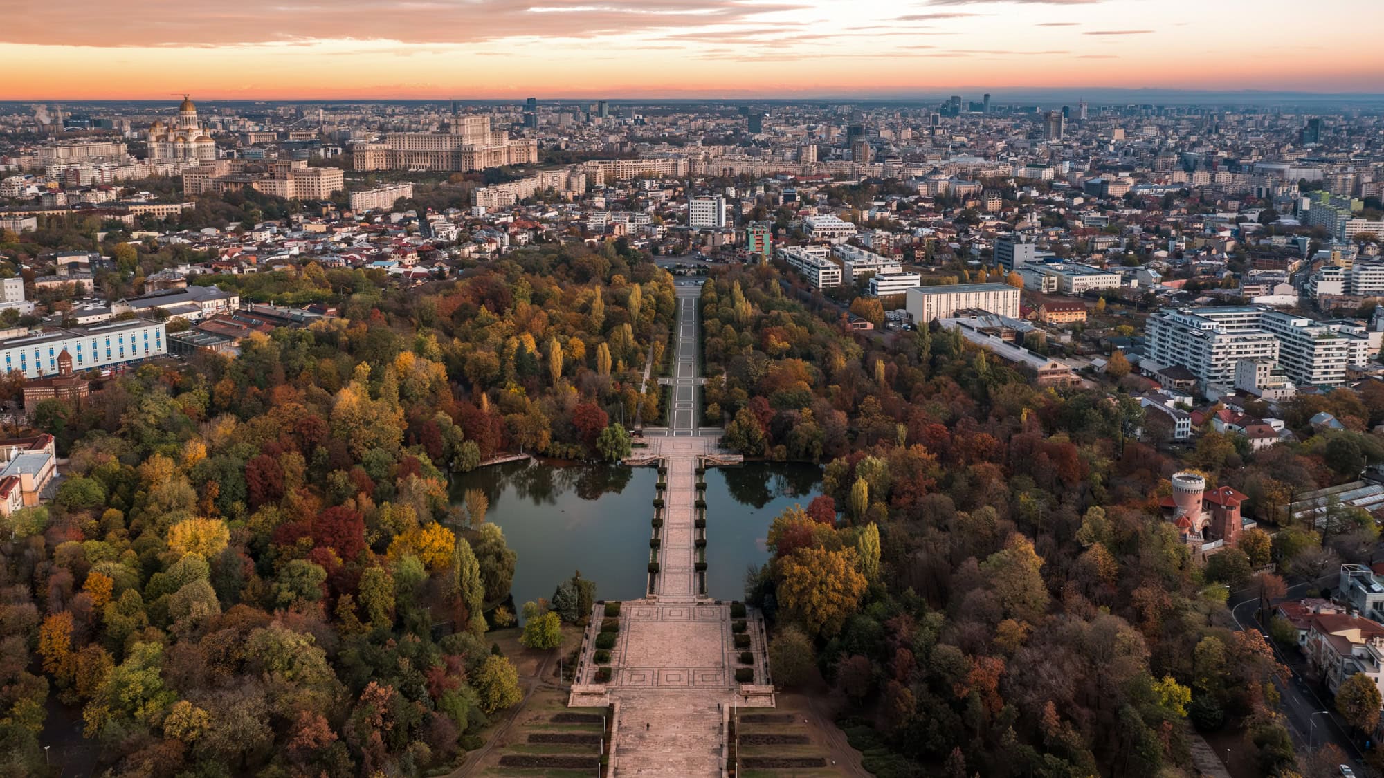 A sweeping aerial view of Bucharest at sunset, featuring the symmetrical layout of Carol Park in vibrant autumn foliage, with its central promenade and reflecting pools leading the eye toward the massive Palace of the Parliament dominating the city skyline in the distance.