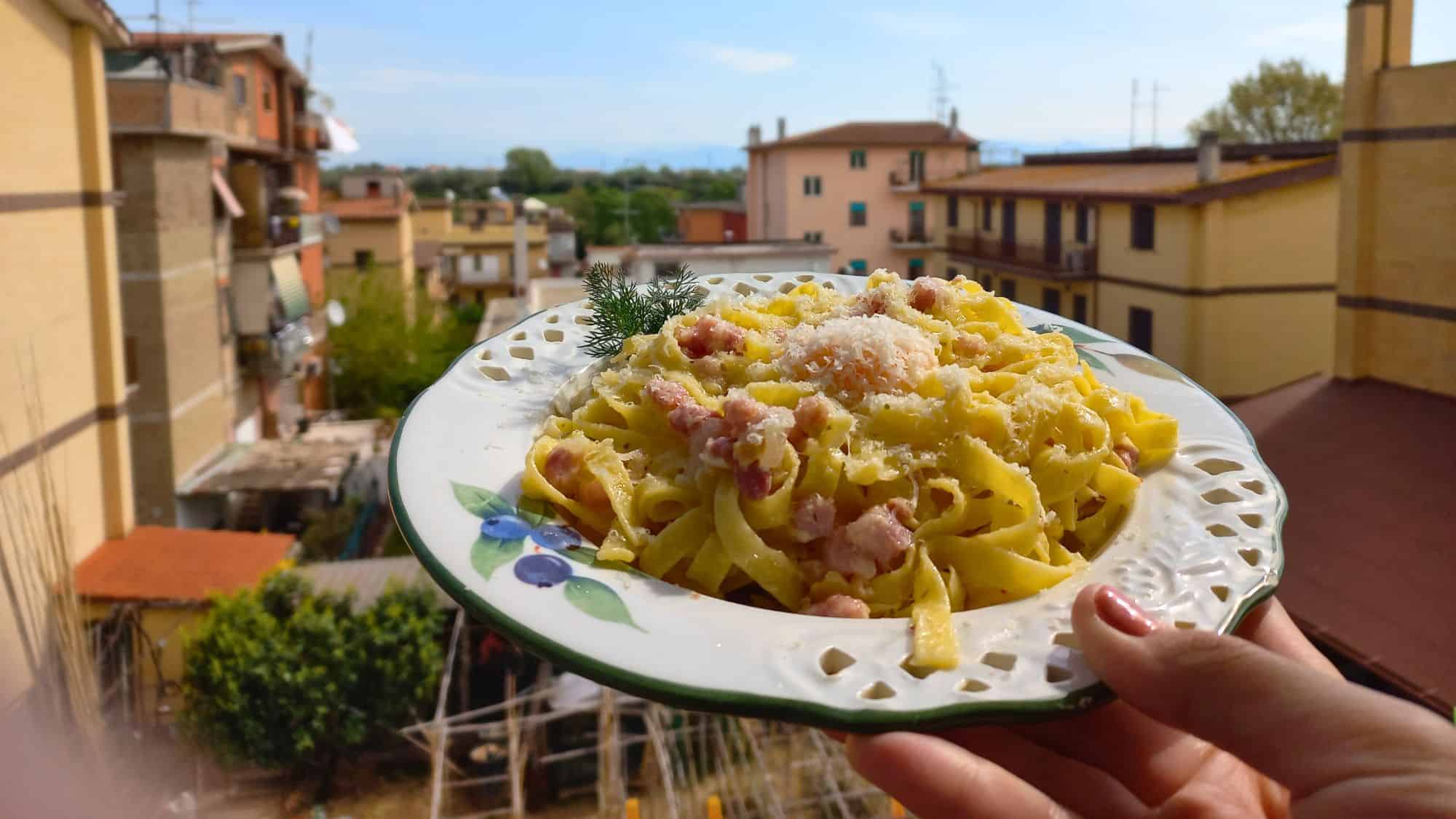 A hand holds a decorative plate of creamy pasta alla carbonara, garnished with pancetta and cheese, against the backdrop of a residential Roman neighborhood.