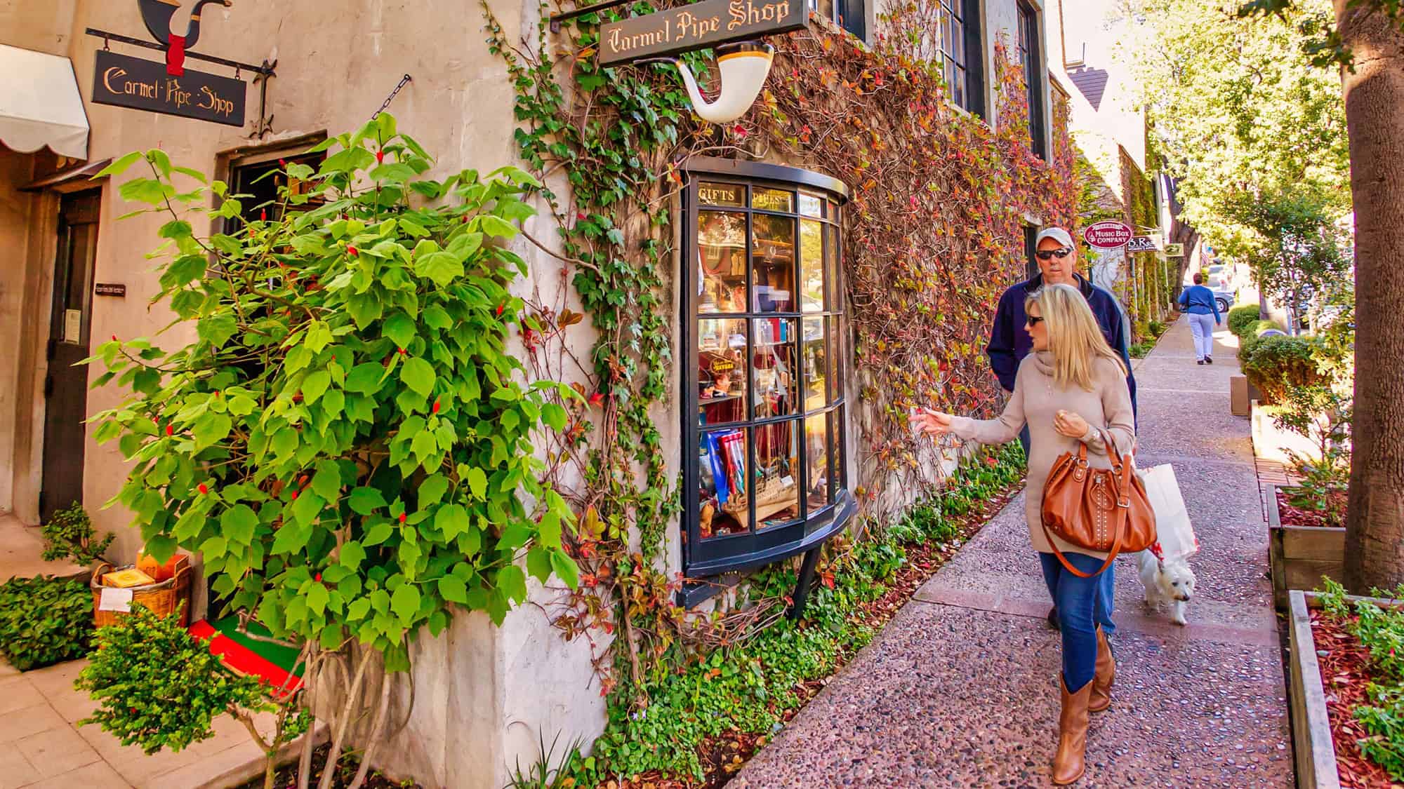 Charming boutique-lined sidewalk in Carmel-by-the-Sea, California, where a woman walks her dog past ivy-covered walls and a vintage-looking "Carmel Pipe Shop" sign.