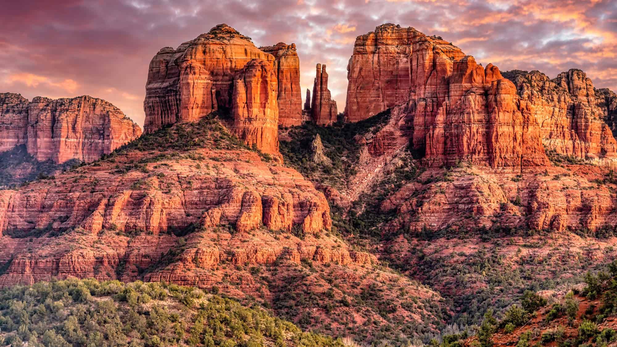 Cathedral Rock’s towering sandstone spires dominate the Sedona skyline under a pink and purple sunset, with forested slopes at their base adding contrast.
