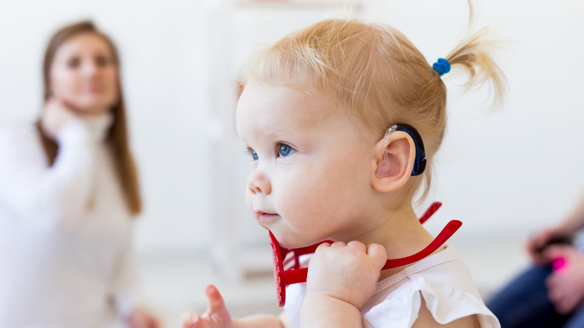 A toddler with blonde hair and a blue hearing aid looks attentively to the side while holding red toy glasses, with adults softly blurred in the background.