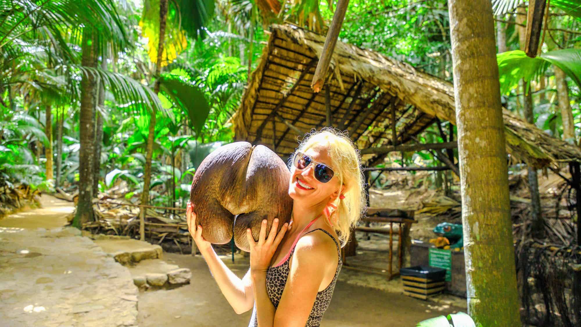 A smiling woman in sunglasses holds a massive Coco de Mer seed with both hands in a lush tropical forest, standing near a rustic thatched hut in Seychelles.