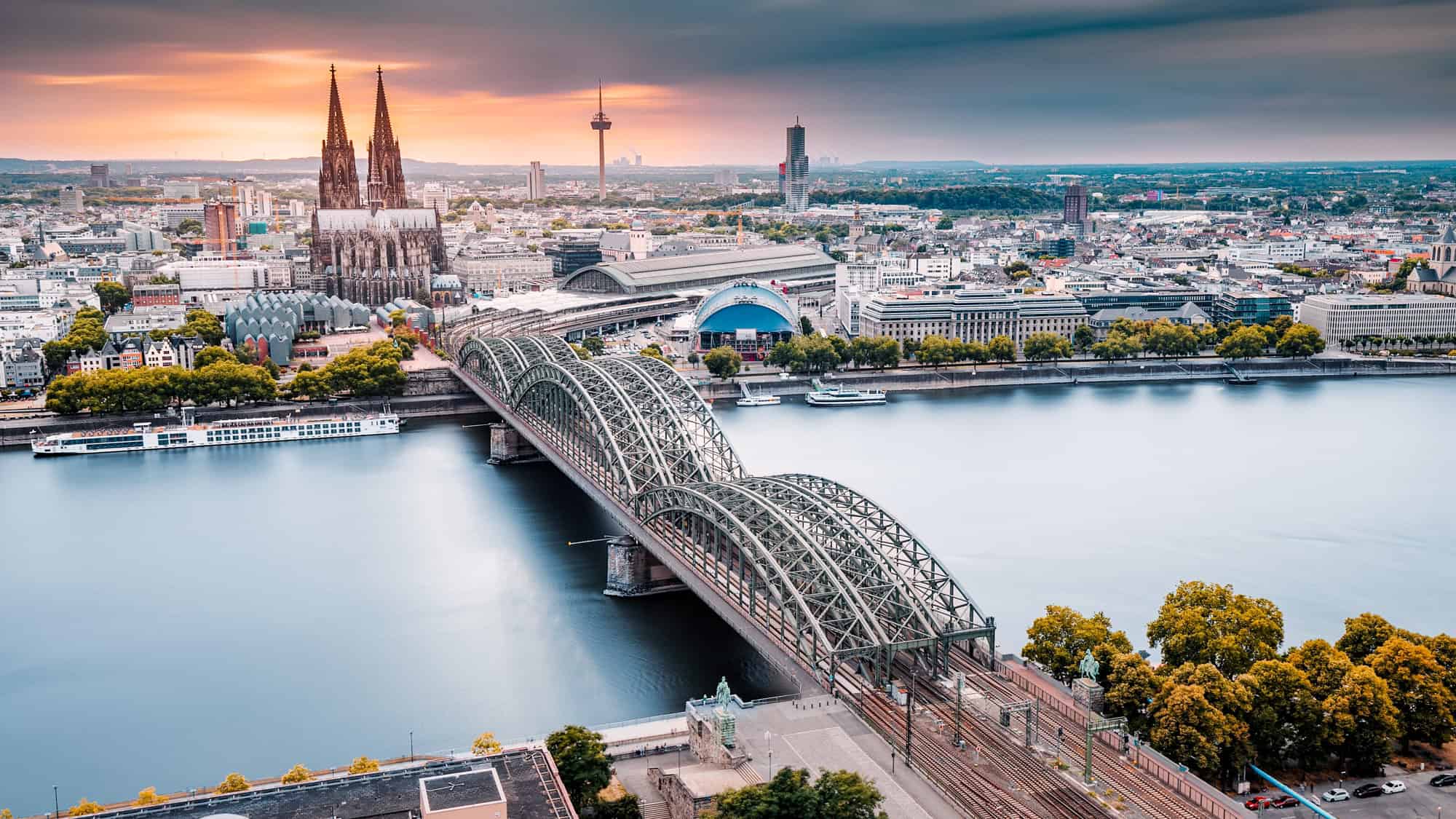 Aerial view of Cologne, Germany, showing the twin spires of Cologne Cathedral, the Hohenzollern Bridge spanning the Rhine River, and the cityscape glowing under a moody sunset sky.