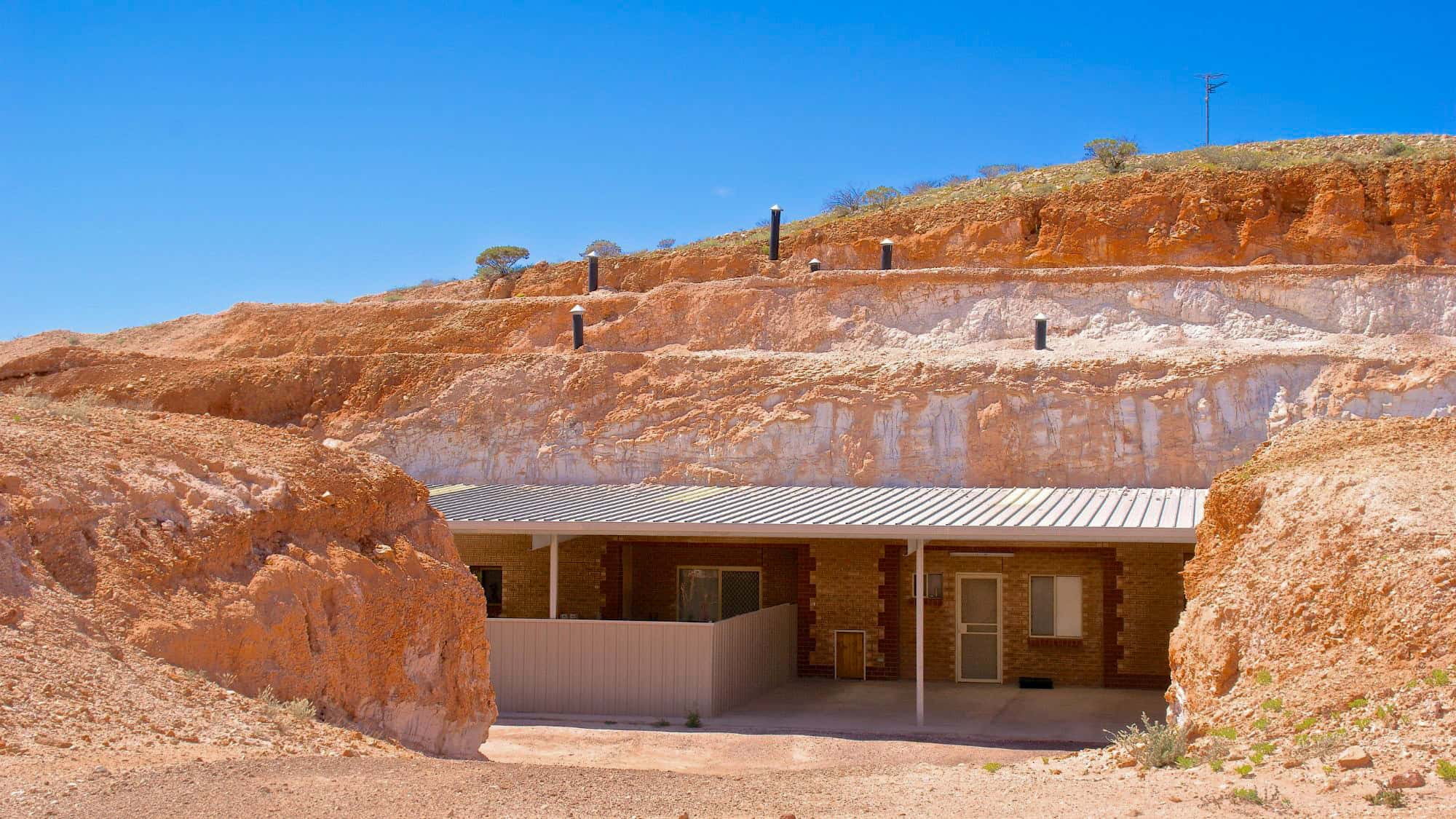 A home built into a rocky hillside in Coober Pedy, with only the front entrance visible and the rest tunneled underground; the terrain is dry and reddish, with ventilation pipes dotting the slope above the structure.