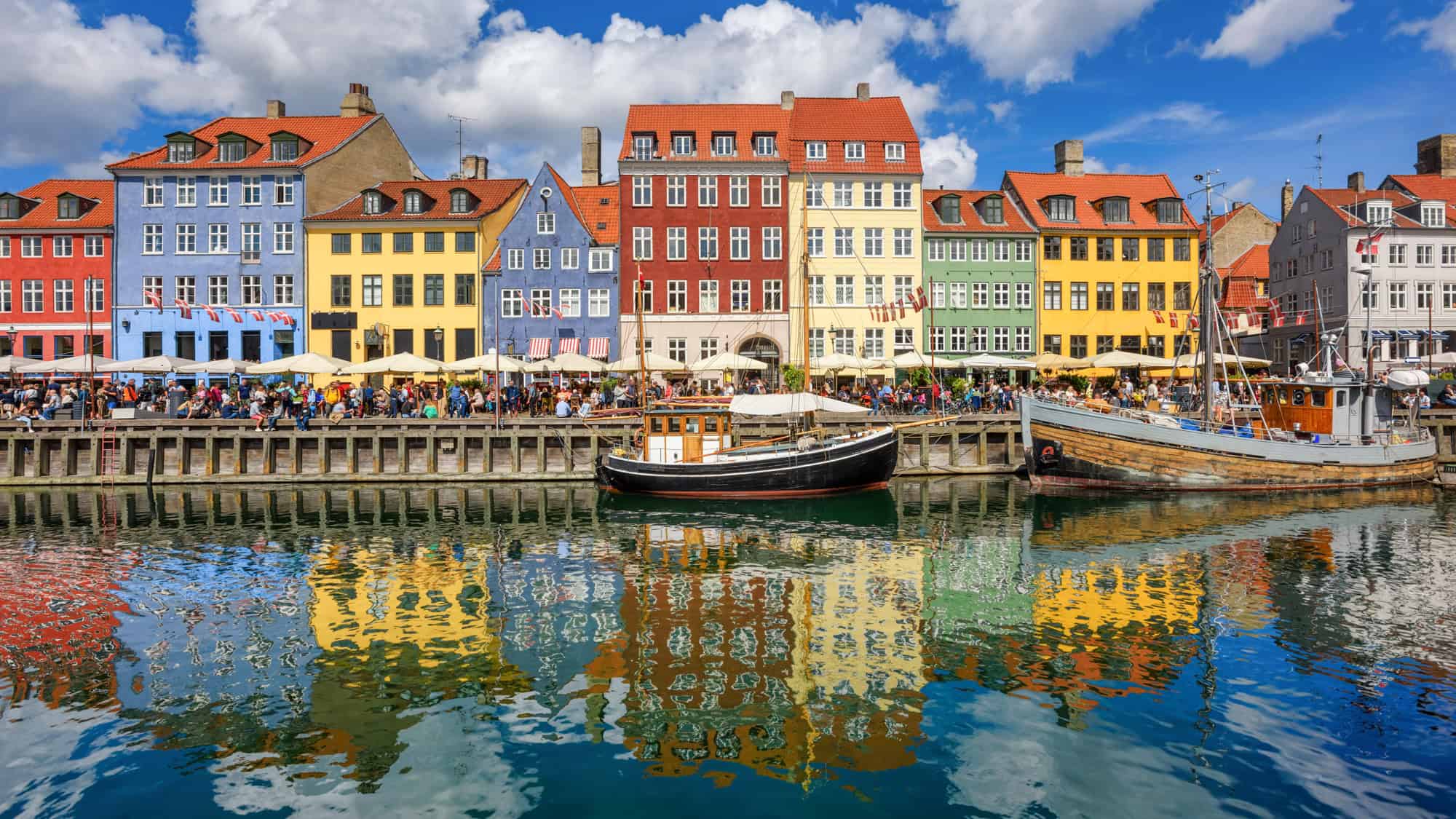 Colorful historic buildings line the waterfront in Copenhagen’s Nyhavn district, with outdoor cafes bustling with people and boats docked in the canal reflecting the vibrant facades.