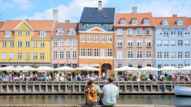 A couple sits by the Nyhavn canal in Copenhagen, Denmark, facing rows of pastel-colored buildings and umbrellas shading diners along the lively waterfront promenade.