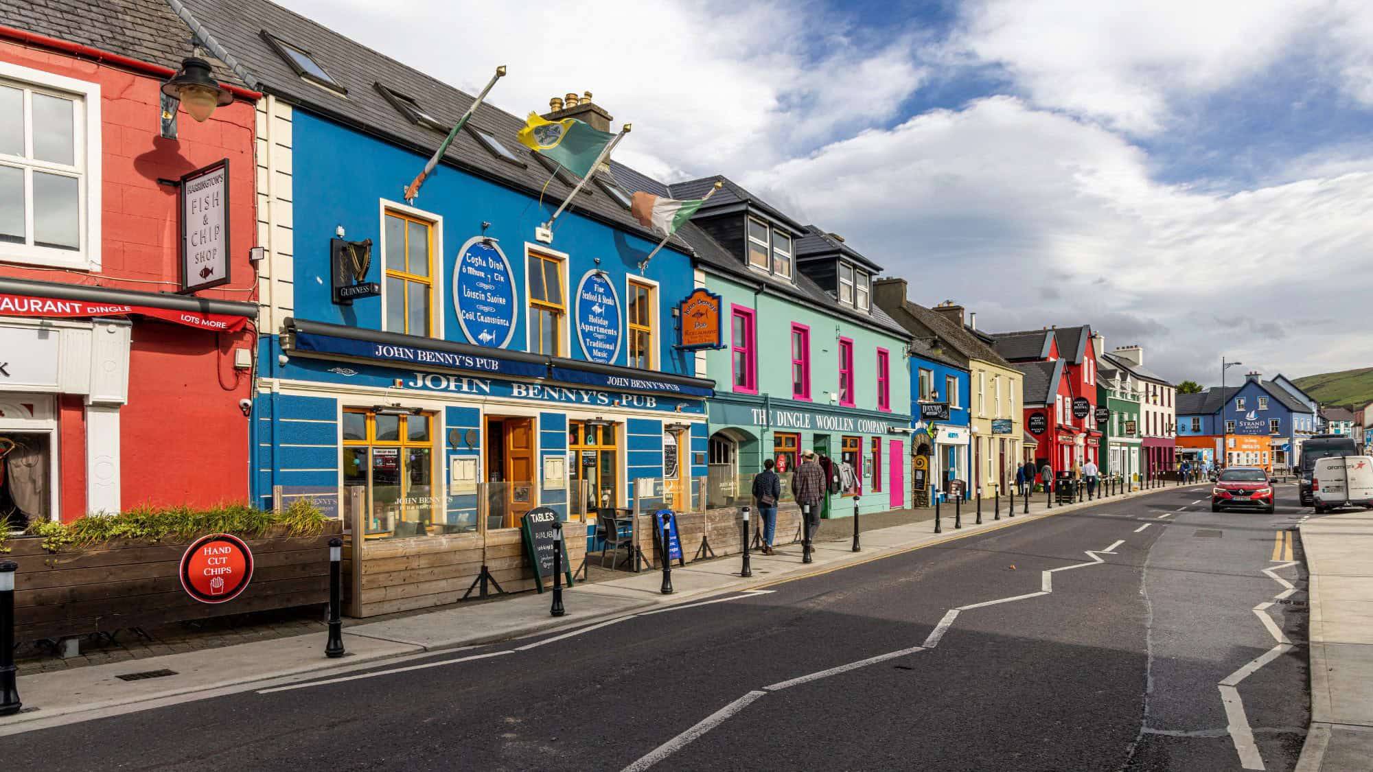 A lively row of brightly painted storefronts and pubs along the main street in Dingle, Ireland, with pedestrians and cars under partly cloudy skies.