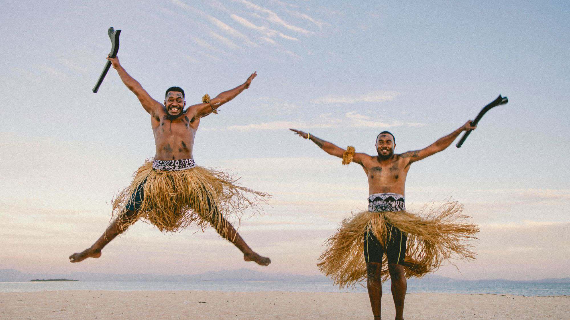 Two men in traditional Fijian attire leap joyfully on a sandy beach, holding wooden clubs and wearing grass skirts, with the ocean and sky behind them.