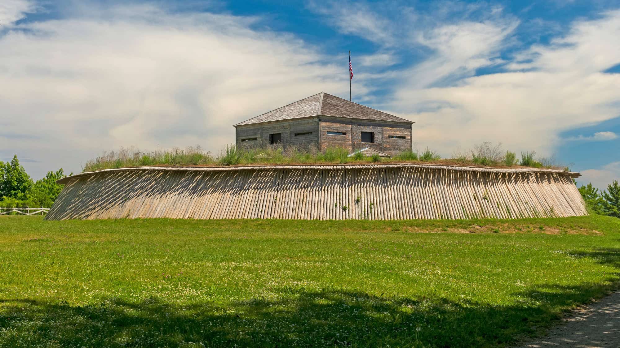 Historic wooden blockhouse of Fort Holmes sits atop a grassy hill surrounded by a slanted wooden stockade, under a partly cloudy sky on Mackinac Island.