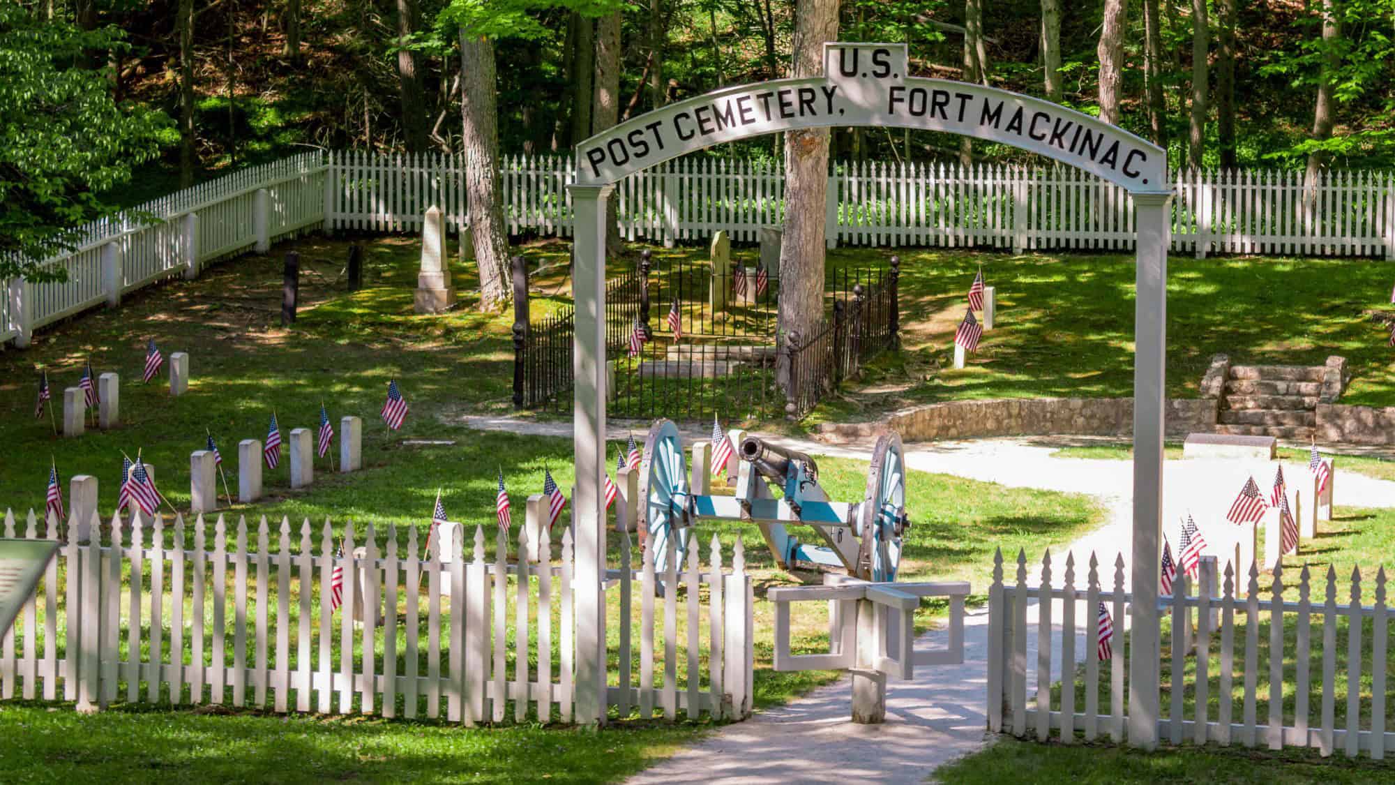 The entrance to Fort Mackinac Post Cemetery features a cannon behind a white picket fence, American flags placed near gravestones, and dappled sunlight filtering through surrounding trees.