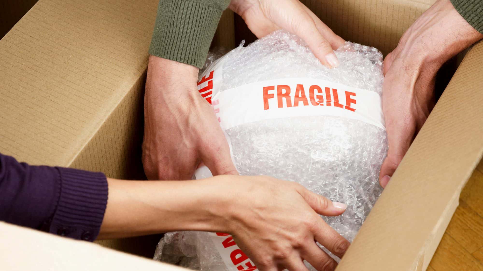 Two pairs of hands gently wrap a large, bubble-wrapped item labeled “FRAGILE” in red text and place it into a brown cardboard moving box.