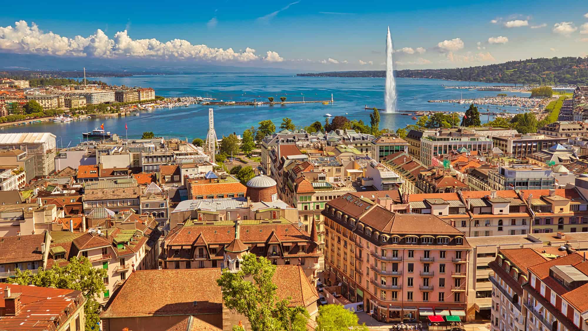 Overhead shot of Geneva, Switzerland, featuring red-tiled rooftops, the shimmering blue waters of Lake Geneva, and the iconic Jet d’Eau fountain spraying high into a clear, sunny sky.