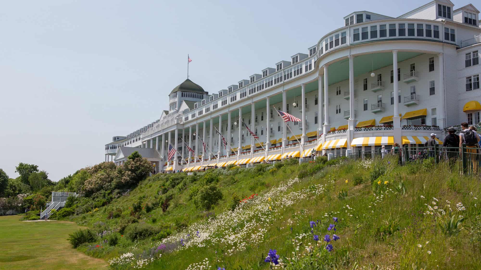 The iconic Grand Hotel on Mackinac Island, with its long white colonnade, American flags, and yellow-striped awnings, overlooking a garden of wildflowers on a sunny day.