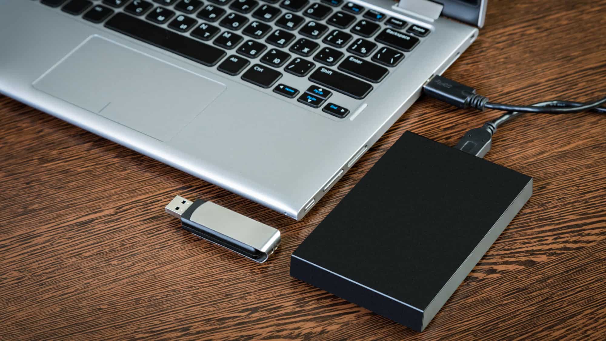 A silver laptop on a wooden table is connected to an external hard drive, with a USB flash drive placed beside it, suggesting data storage or backup.