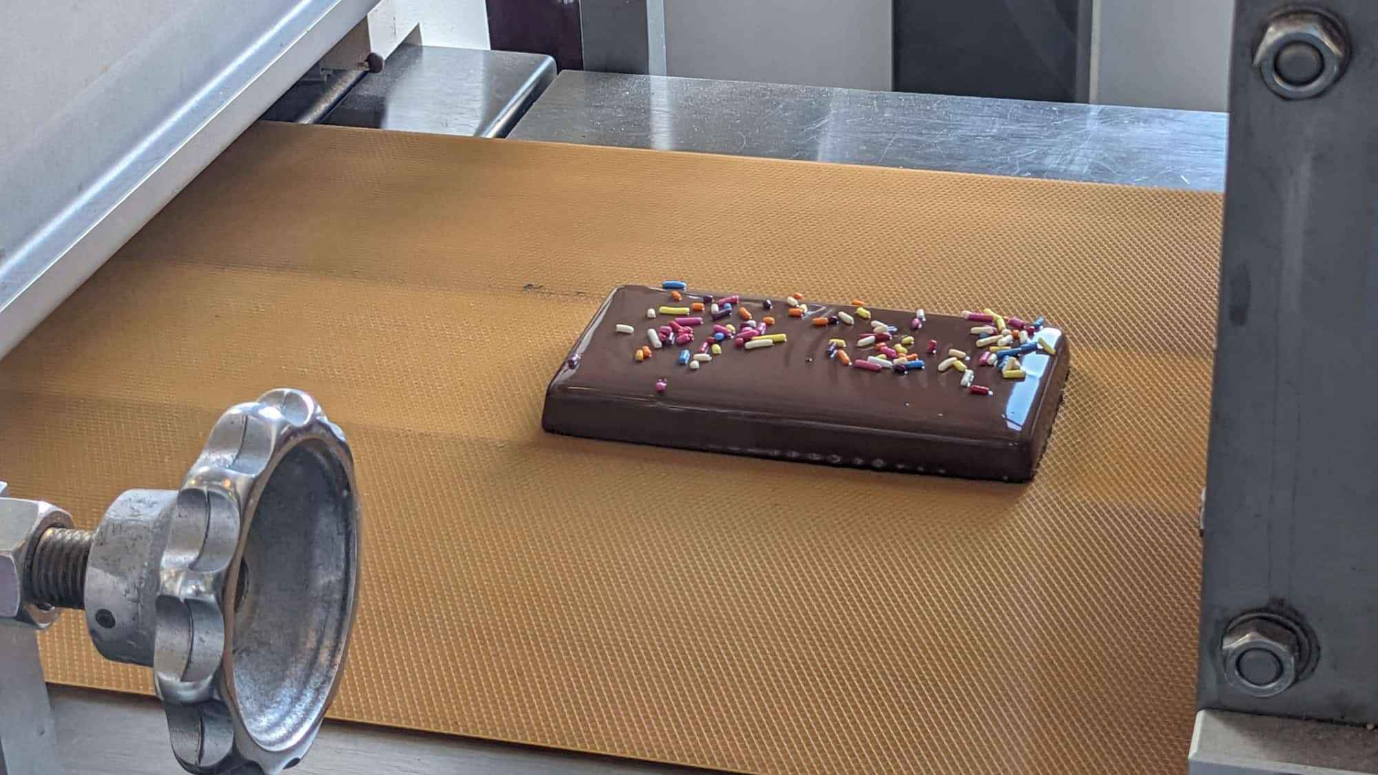 A single chocolate bar topped with colorful sprinkles rides a yellow conveyor belt inside a factory, likely part of a chocolate-making process at Hershey.