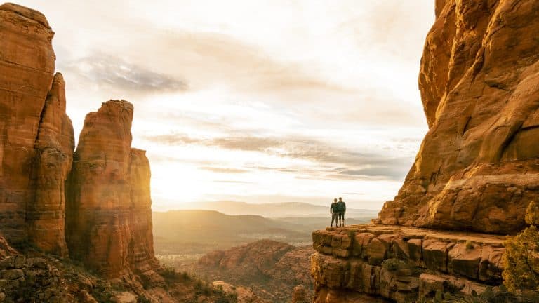 Two people stand atop a dramatic red rock cliff in Sedona at golden hour, dwarfed by the towering formations around them and bathed in warm sunlight.