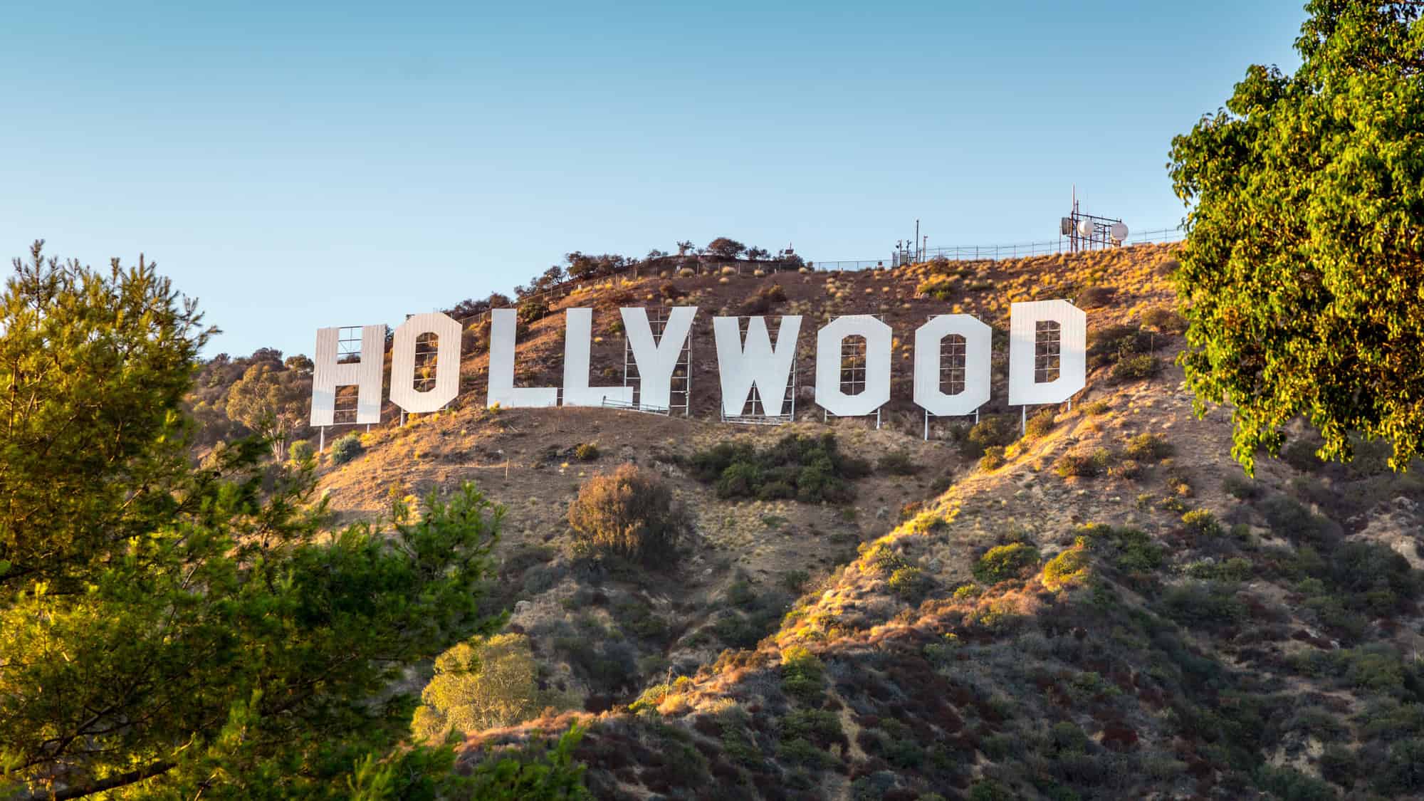 Iconic image of the large white “HOLLYWOOD” sign perched on the dry, rugged hillside of the Hollywood Hills in Los Angeles, California. The surrounding terrain is dotted with bushes and trees under a clear blue sky.