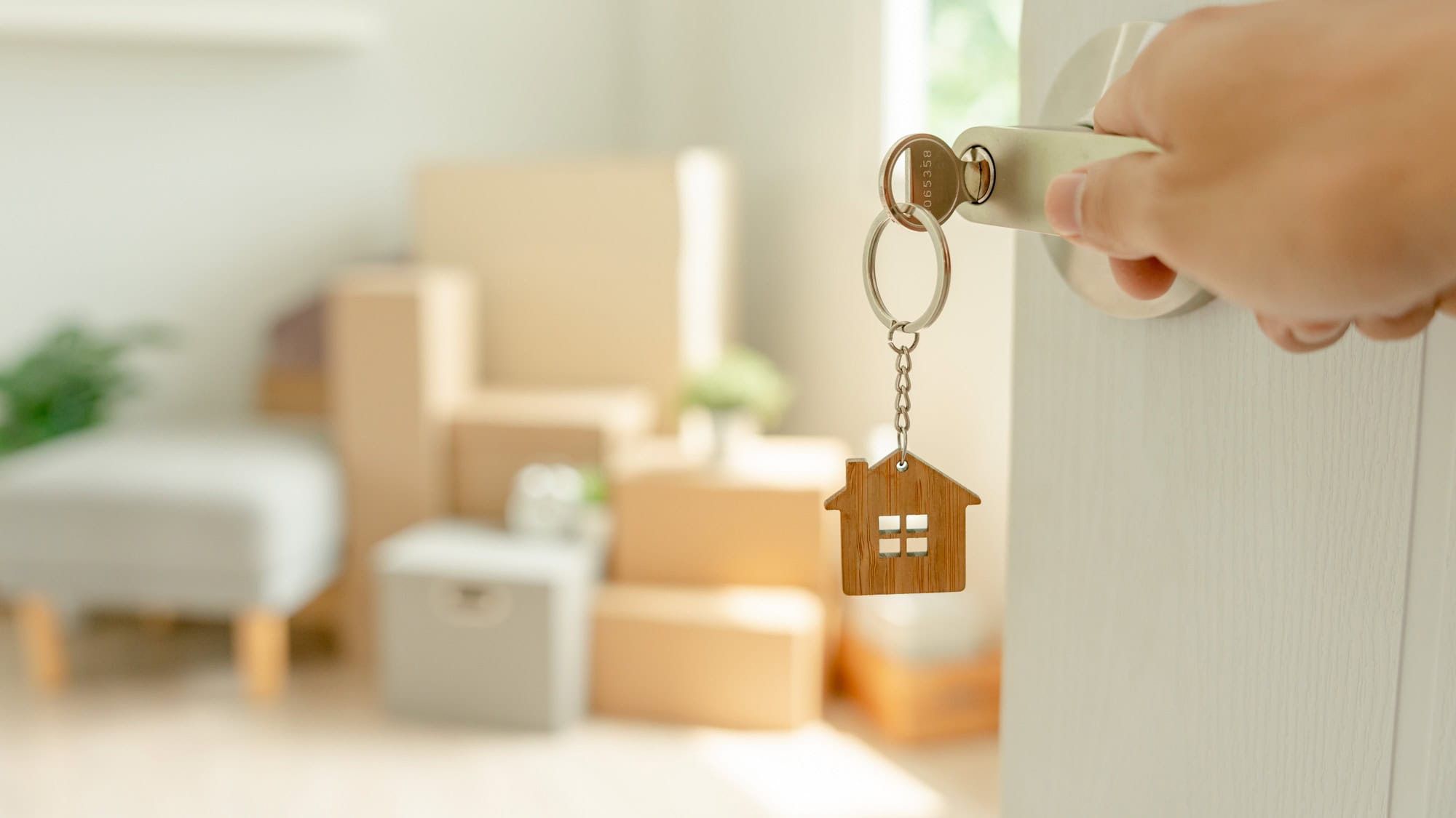 A hand holds a key inserted in a white door, with a wooden house-shaped keychain dangling, and cardboard boxes are seen stacked in the softly blurred background.