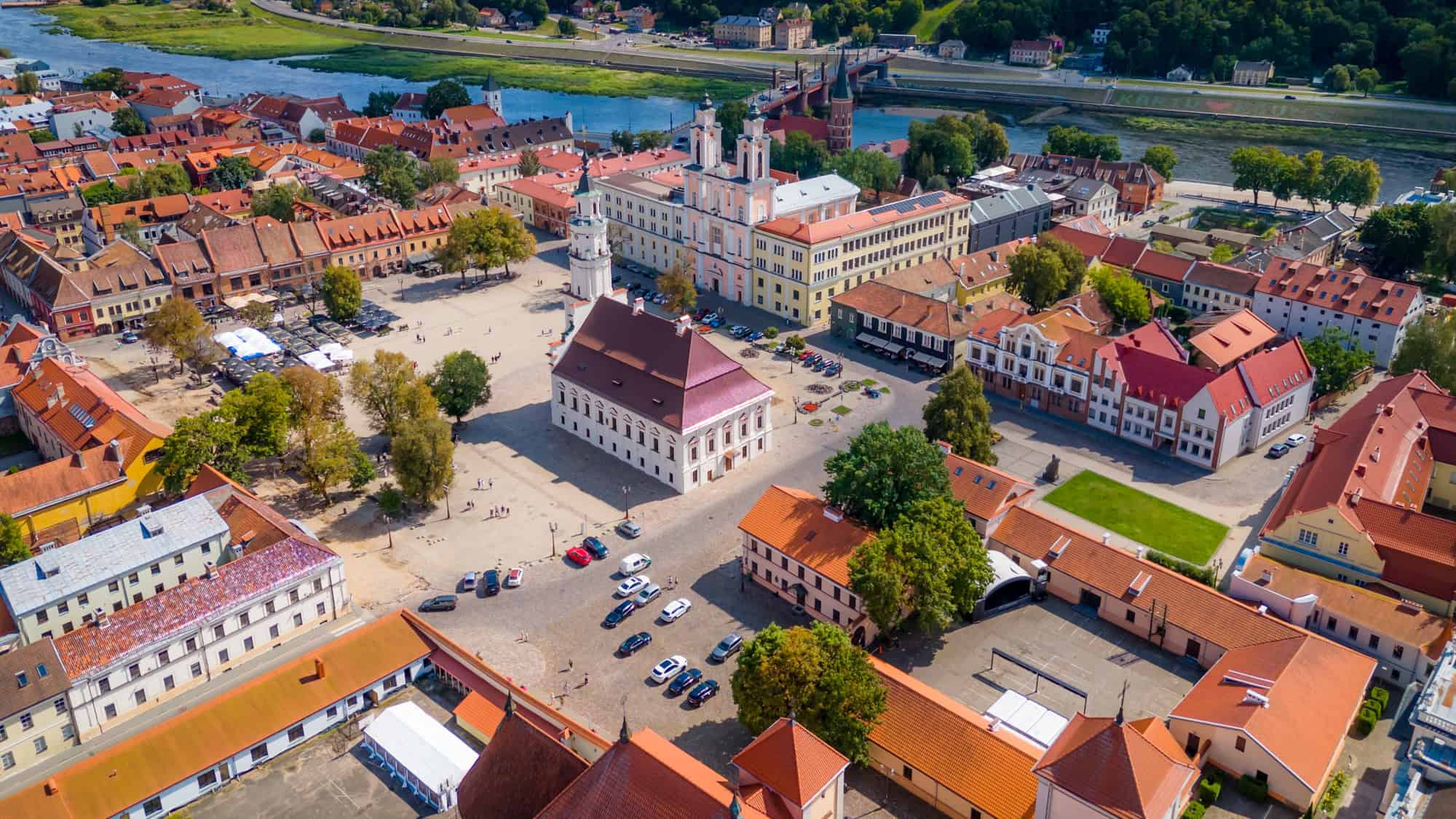 Aerial view of the Kaunas Town Hall Square, with its elegant central white town hall building surrounded by red-roofed historic homes, a river in the distance, and a quaint, orderly layout of cobbled streets.