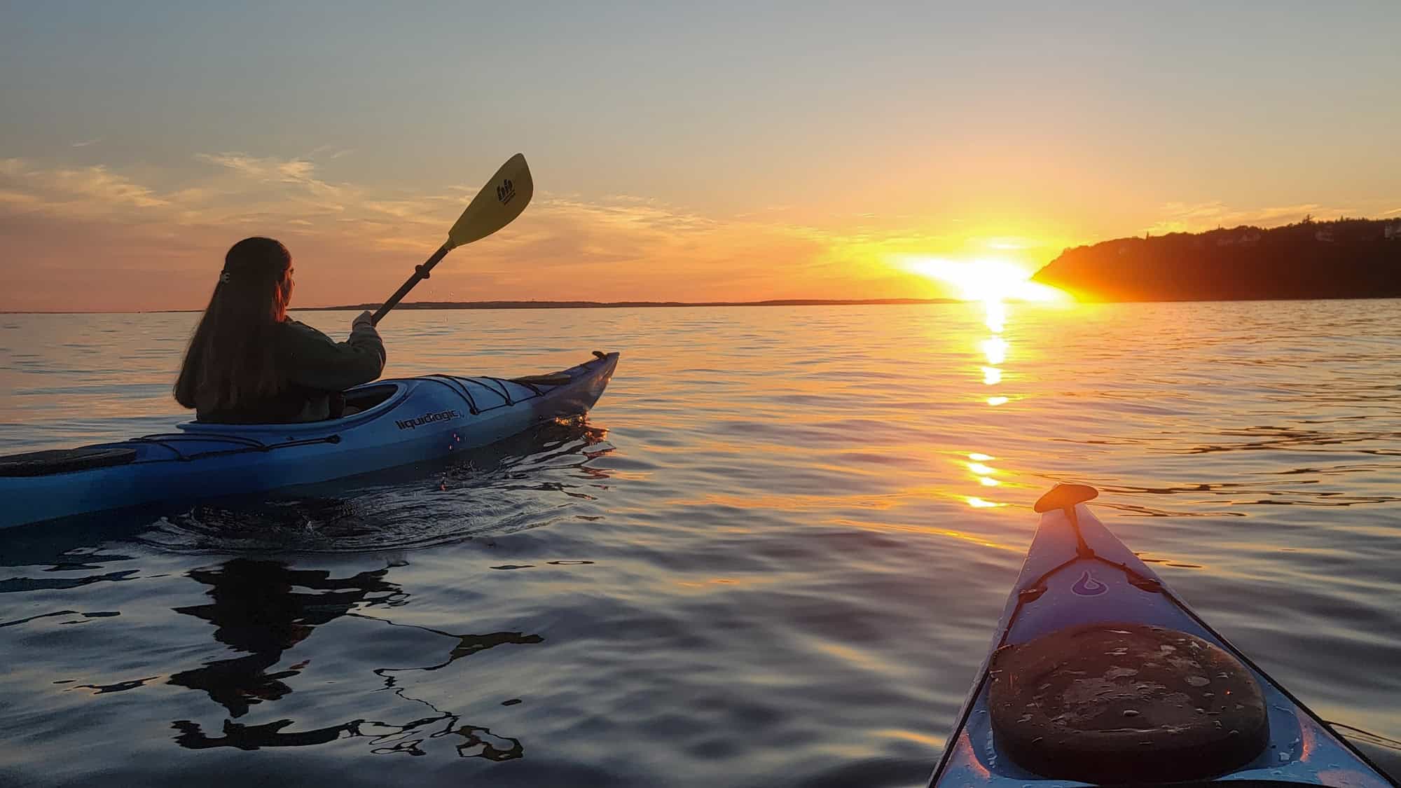 A woman paddles a blue kayak across calm lake waters at sunset, with the golden sun low on the horizon and another kayak’s bow in the foreground.