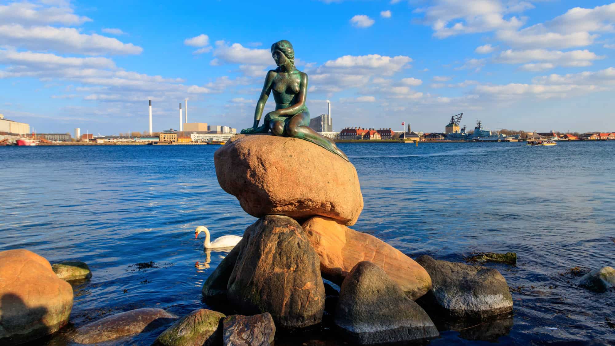 A bronze statue of a mermaid sits gracefully on a rock by the water’s edge in Copenhagen. The cityscape and a swan in the harbor provide a serene backdrop to this tribute to Hans Christian Andersen’s fairytale.