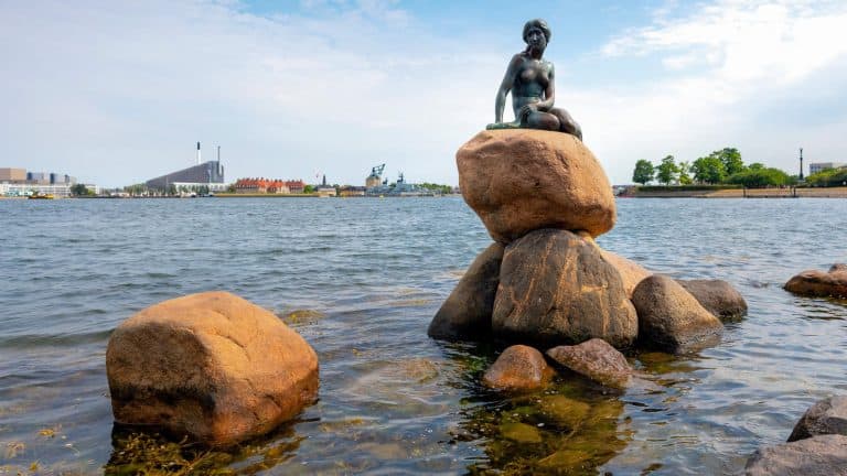Photograph of the famous Little Mermaid statue in Copenhagen, Denmark, showing the bronze mermaid perched on a rock by the water’s edge with a clearer view of the cityscape and modern architecture in the background. This version is taken from a slightly different angle and lighting than the earlier image.