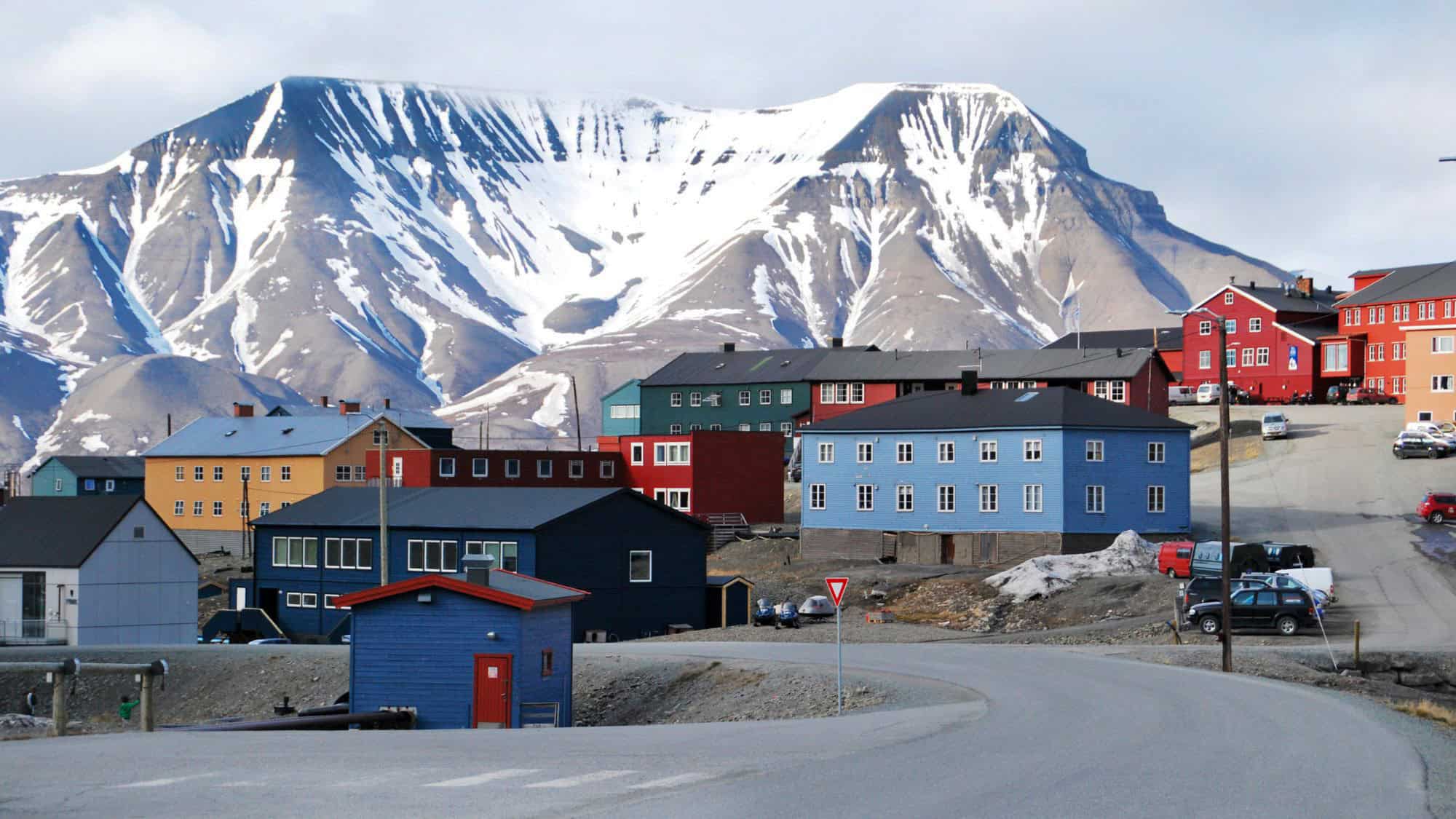 A colorful Arctic town with red, blue, yellow, and green buildings standing out against snow-streaked mountains, under an overcast sky.