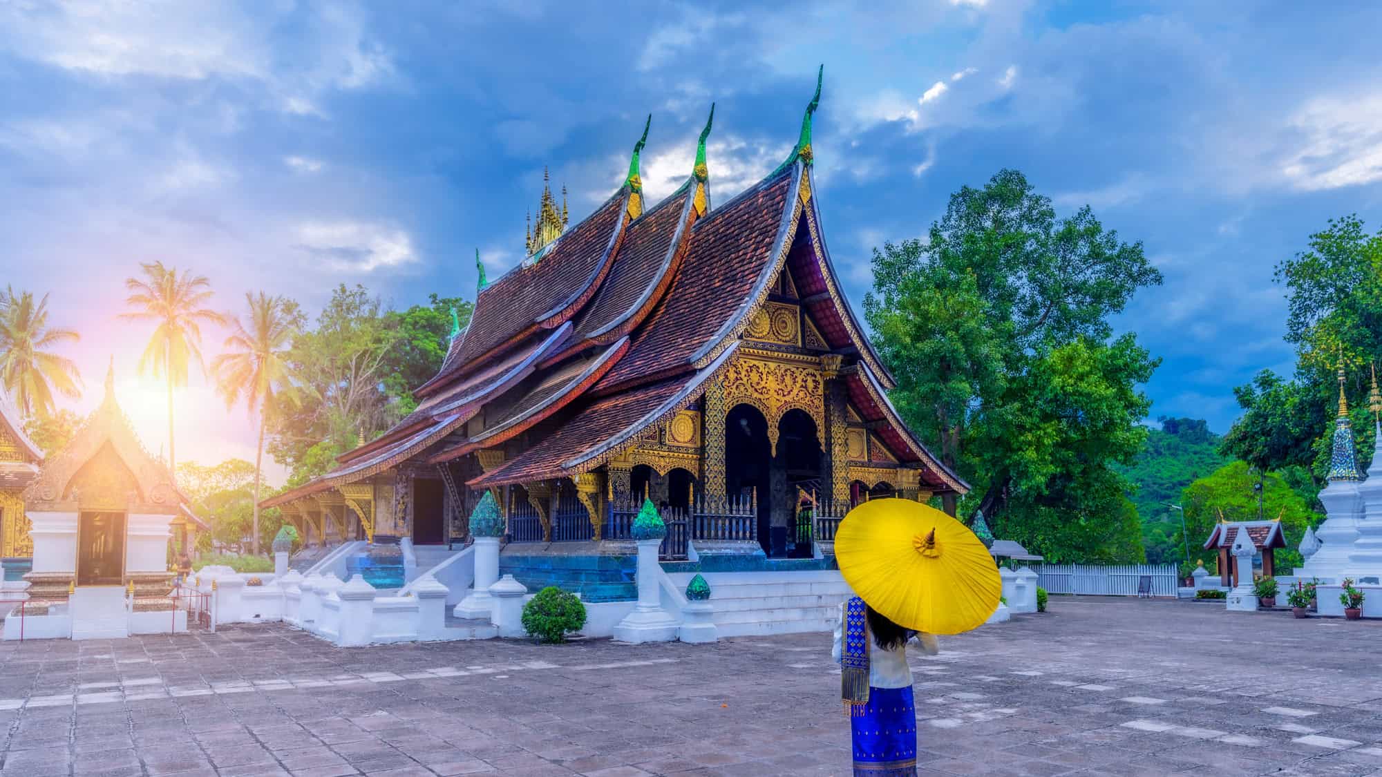 A woman in traditional Lao attire with a yellow parasol stands in front of the elaborately decorated Wat Xieng Thong temple in Luang Prabang, Laos, as the sun sets behind lush tropical trees.