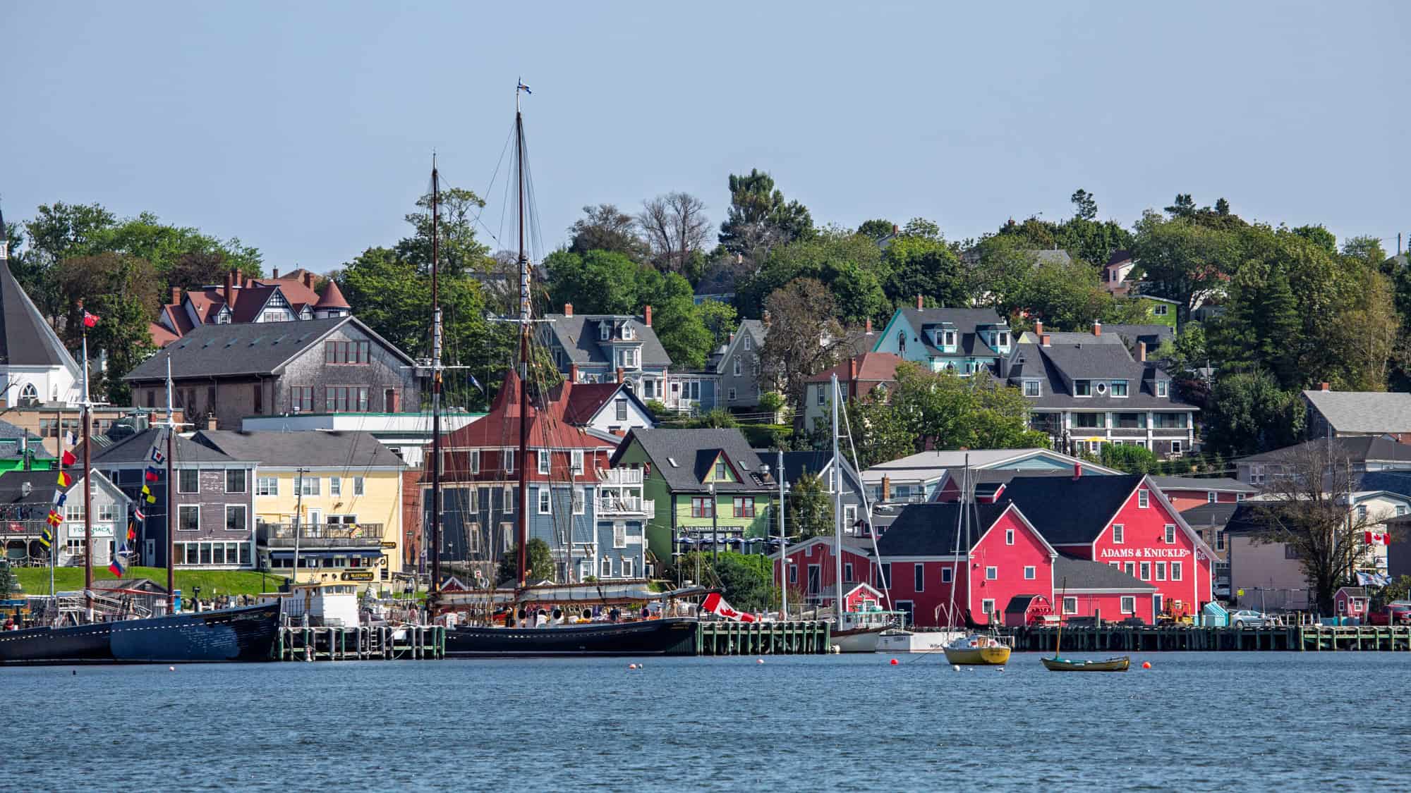 Colorful historic buildings and sailing ships line the waterfront in Lunenburg, Nova Scotia, with Victorian-style homes rising behind the harbor on a clear summer day.