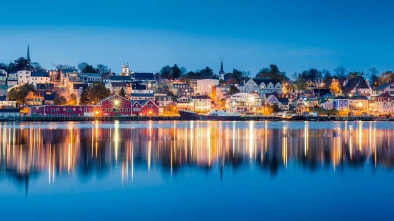 A scenic waterfront view of Lunenburg, Nova Scotia, taken at dusk, reflects the town's colorful buildings and glowing lights across still water. The iconic red buildings of the harbor and the church steeples add historic charm to this UNESCO World Heritage town.