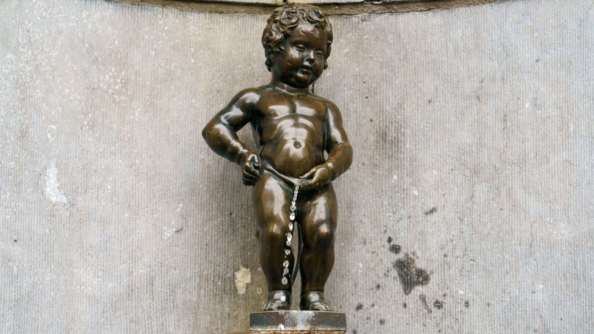 A small bronze statue of a naked boy urinating into a fountain, mounted on a stone wall. Known as Manneken Pis in Brussels, this humorous and iconic statue captures whimsical charm in a compact form.