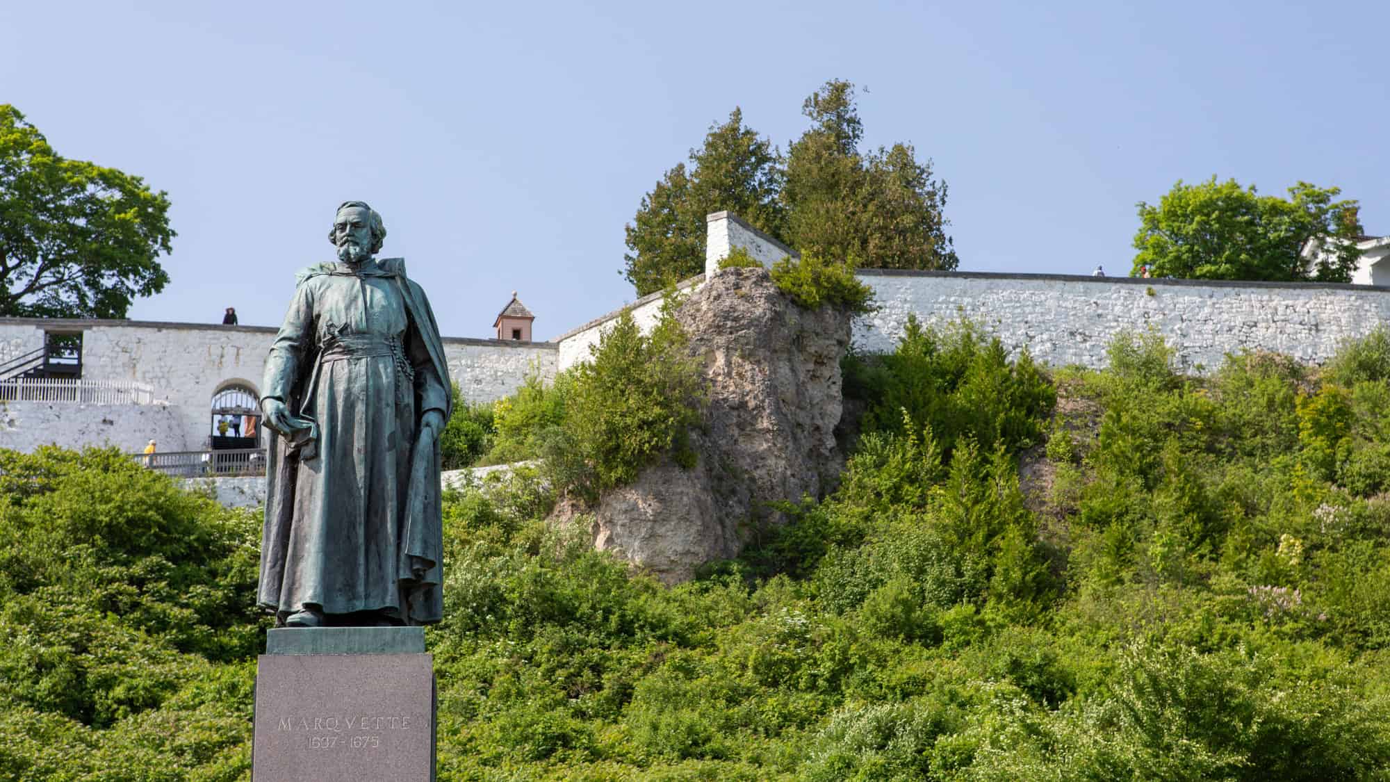 Statue of Father Marquette stands on a stone pedestal in front of a lush hillside and historic white fort walls at Marquette Park on Mackinac Island.