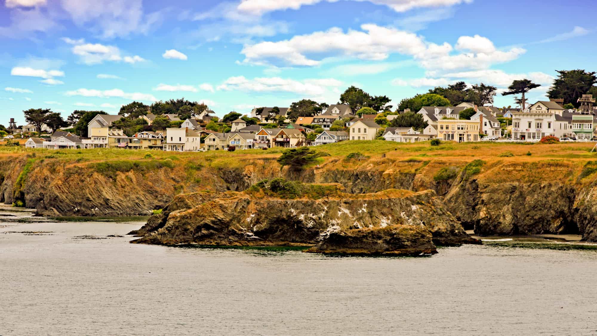 Charming coastal village perched on golden cliffs above the ocean, featuring historic white and pastel homes with gabled roofs, surrounded by a blue sky and soft clouds.