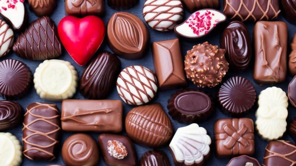 Flat lay of an assorted collection of artisanal chocolates in various shapes and colors, including dark, milk, and white chocolate pieces, with some decorated with drizzle, nuts, or a bright red heart-shaped candy.