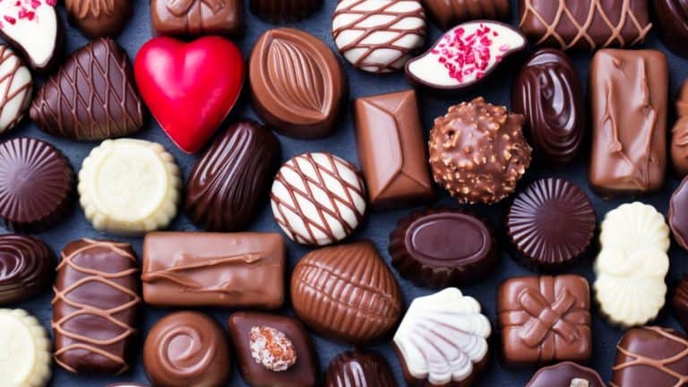 Flat lay of an assorted collection of artisanal chocolates in various shapes and colors, including dark, milk, and white chocolate pieces, with some decorated with drizzle, nuts, or a bright red heart-shaped candy.