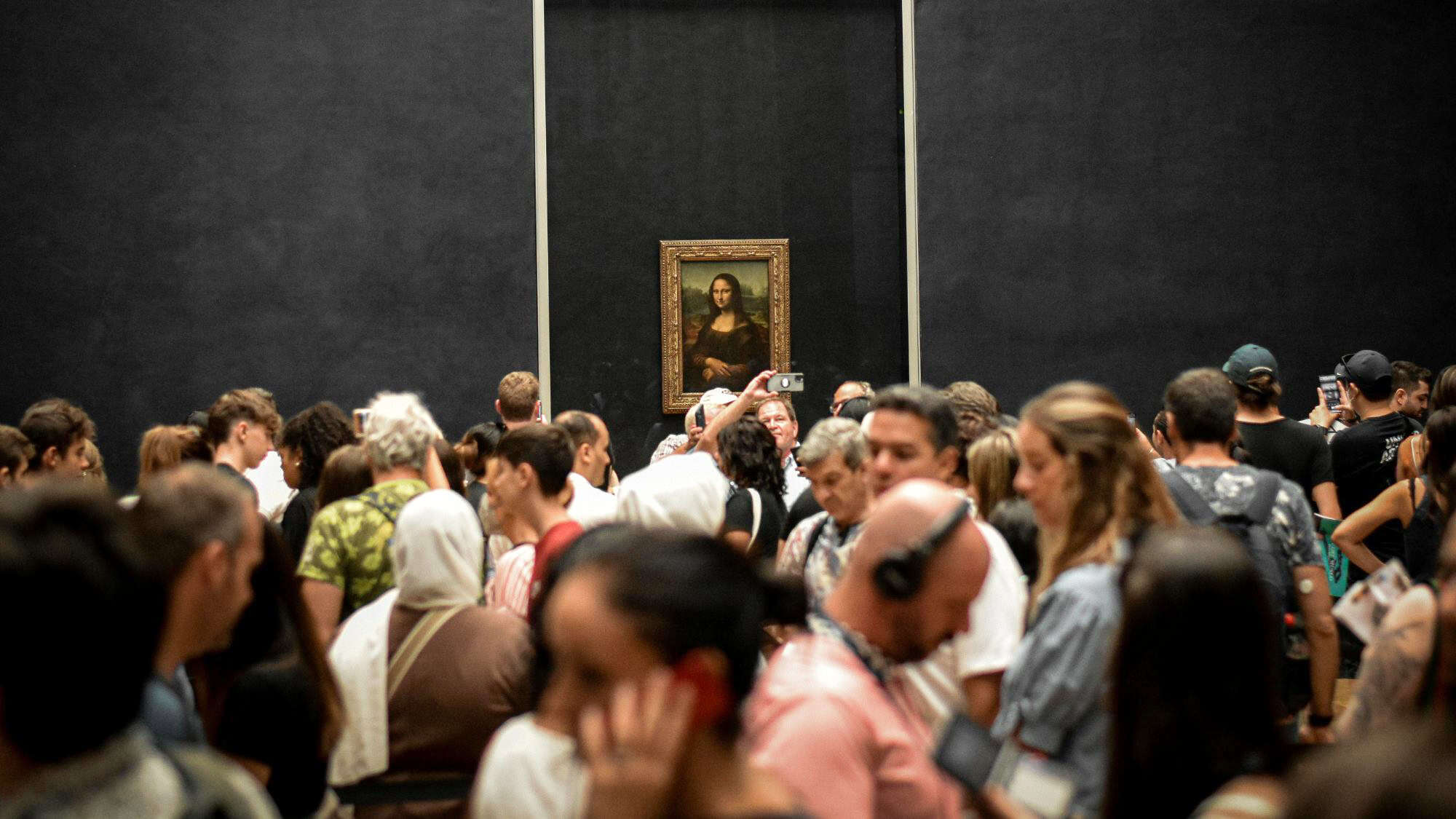 A large crowd gathers in front of the famous Mona Lisa painting at the Louvre Museum in Paris. The painting, displayed behind glass on a dark wall, draws phones and cameras as visitors try to capture a glimpse of Leonardo da Vinci's masterpiece.