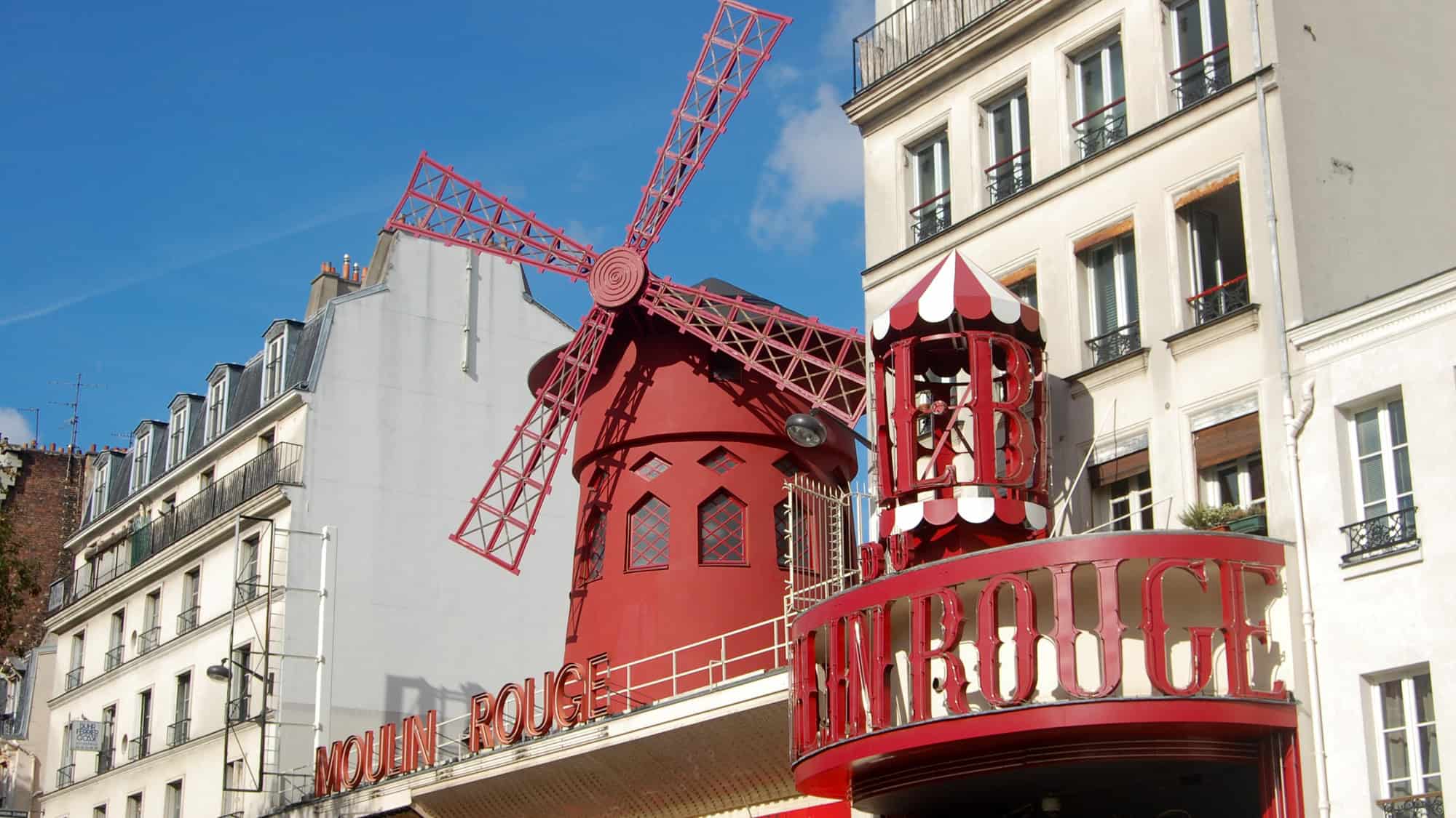 The iconic red windmill of the Moulin Rouge cabaret in Paris stands out against neighboring cream-colored buildings and a bright blue sky. Below the windmill, bold red signage spells out “Moulin Rouge” in vintage-style lettering.