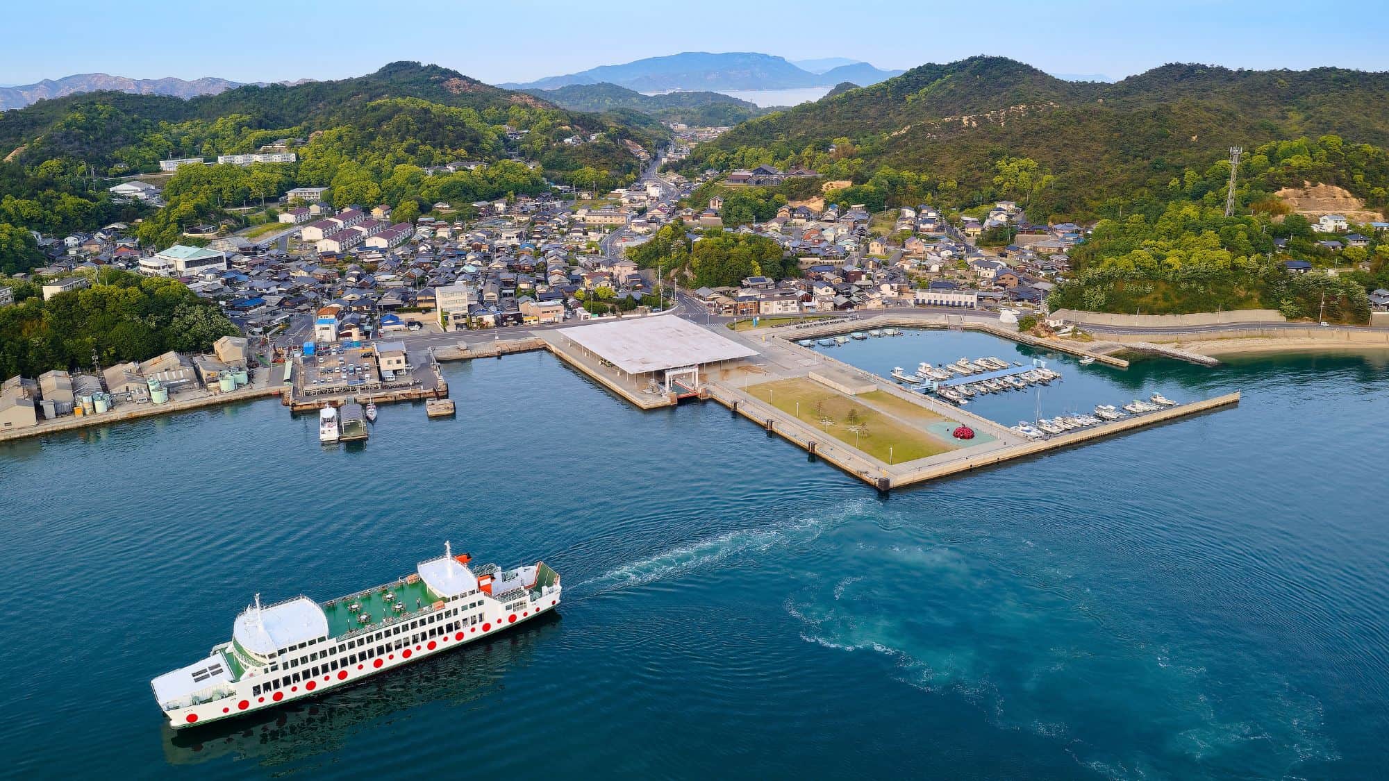 A white ferry with red dots departs a modern dock in Naoshima, Japan, with a view of a compact coastal town surrounded by lush green hills and forested mountains in the background.