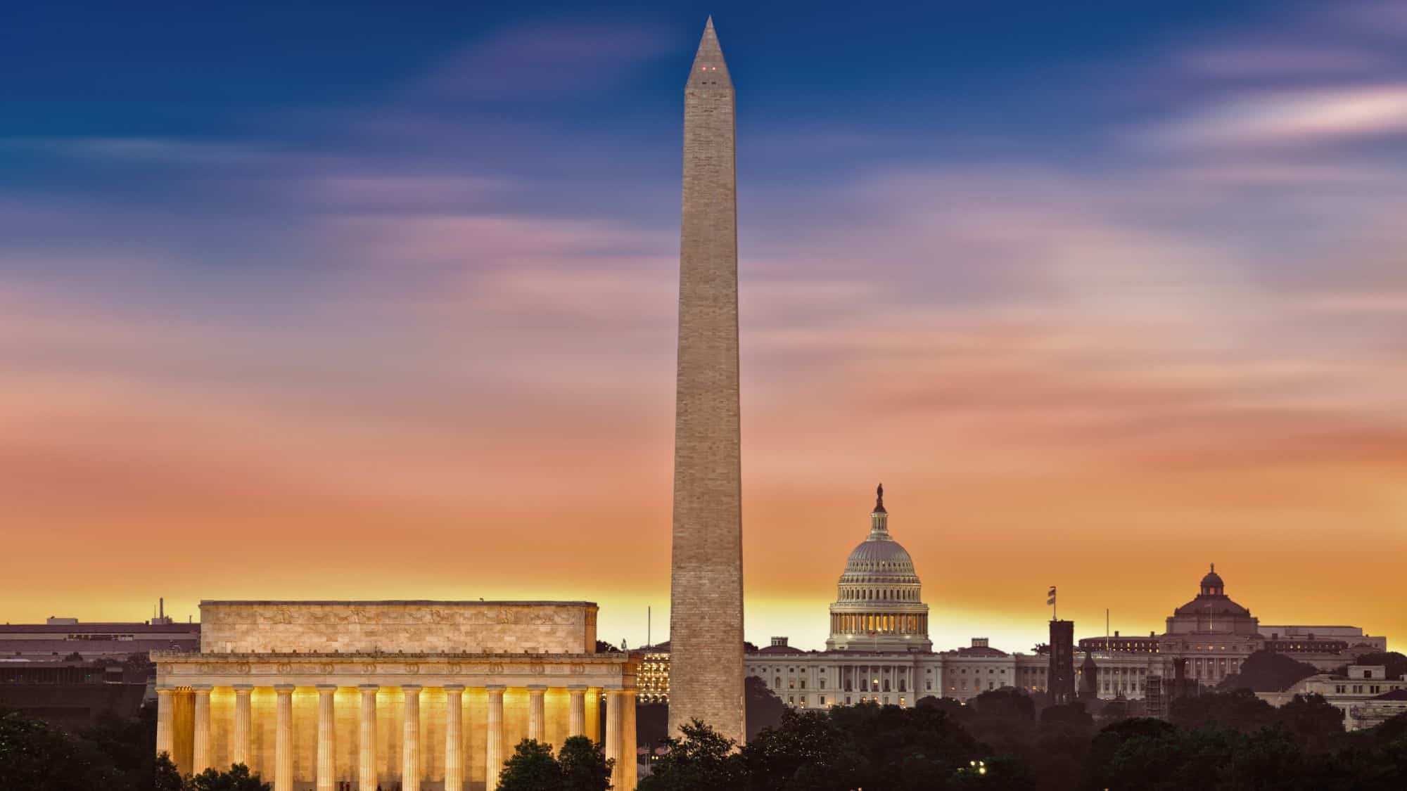 A sunset view of Washington, D.C.’s National Mall with the Washington Monument in the center, flanked by the Lincoln Memorial and U.S. Capitol, glowing under a gradient sky.