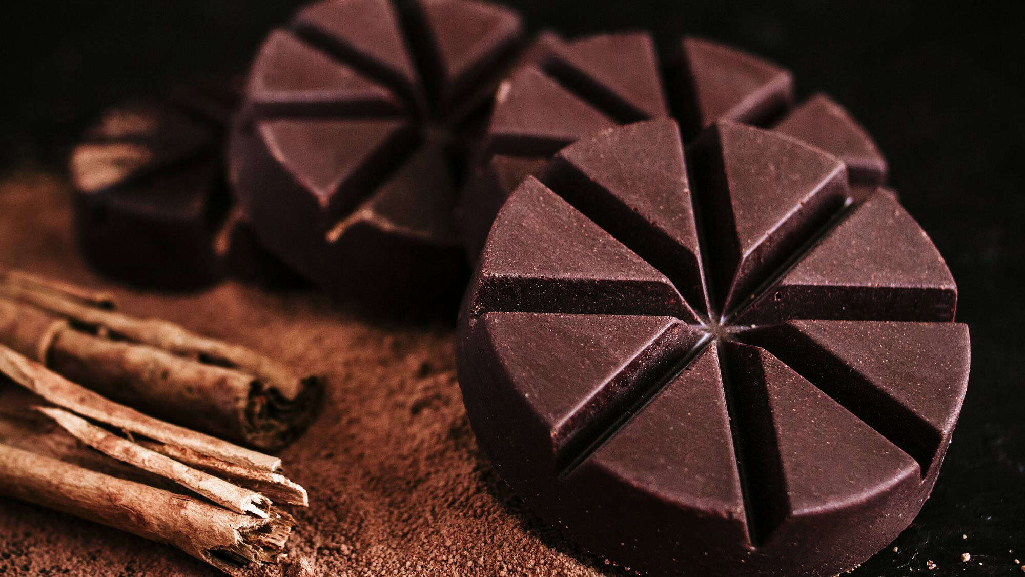 Close-up of rustic, round Mexican chocolate discs with deep grooves, placed on a bed of cocoa powder with cinnamon sticks alongside—evoking rich traditional flavors.