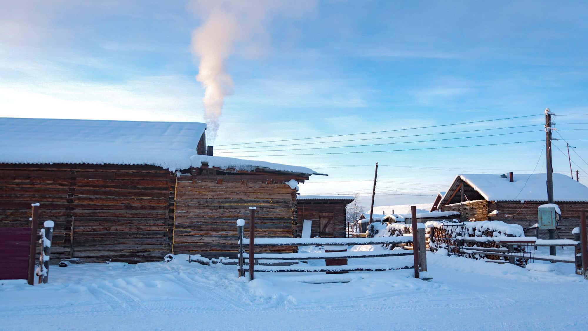 A rustic Siberian village in deep winter, with wooden log cabins blanketed in snow and smoke rising from a chimney into a frosty blue sky.